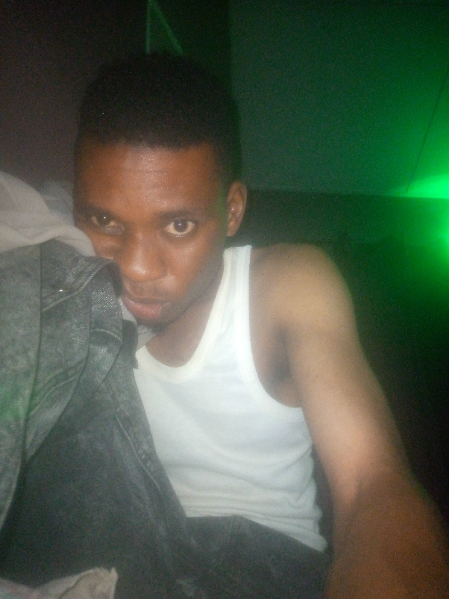 A medium shot shows a man wearing a white tank top, holding a gray denim jacket, possibly indoors, in a location with dark green background lighting. His facial expression is neutral as he looks towards the camera. The location is in the city of Unknown, Unknown.
