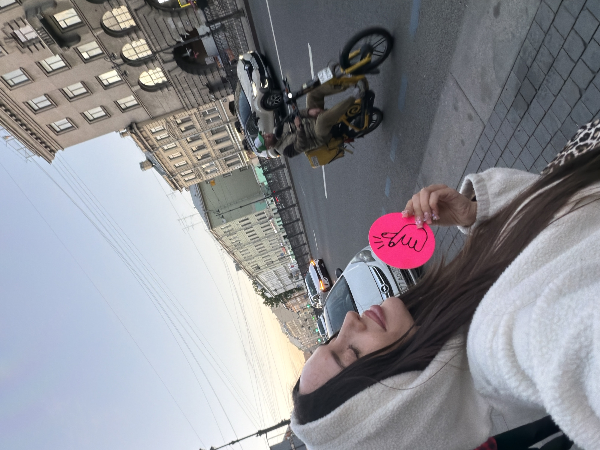 A woman is taking a selfie on a street in Saint Petersburg, Russia, with a pink heart-shaped gesture in her hand and a fluffy white hoodie on. In the background, a delivery person on a yellow scooter is riding down the street, and cars are driving on the road. The buildings in the background are typical of Saint Petersburg architecture. The sky is clear and the sun is setting, casting a warm glow over the scene.