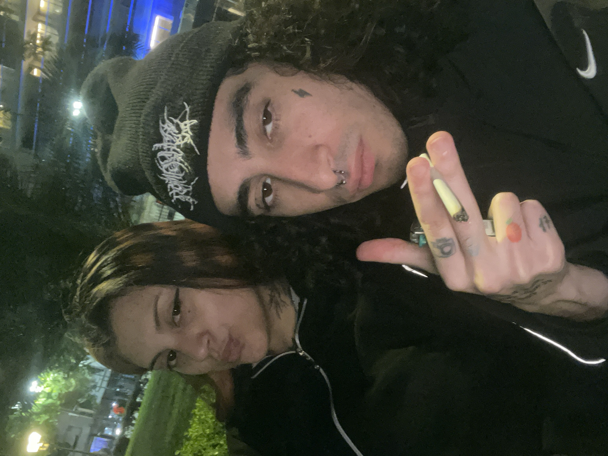 A medium shot captures two individuals in an urban environment. The first, a male with dark curly hair, a tattoo near his left eye, and a nose ring, wears a dark beanie with white lettering and a dark top with a white Nike logo on the right chest. He holds a lit cigarette between his fingers, which are adorned with tattoos. The second individual, a female with short brown hair, wears a dark zip-up jacket with a white stripe. The backdrop shows city lights and trees, suggesting an outdoor setting at night. It is not possible to determine the location in Buenos Aires, Argentina, from the image alone.