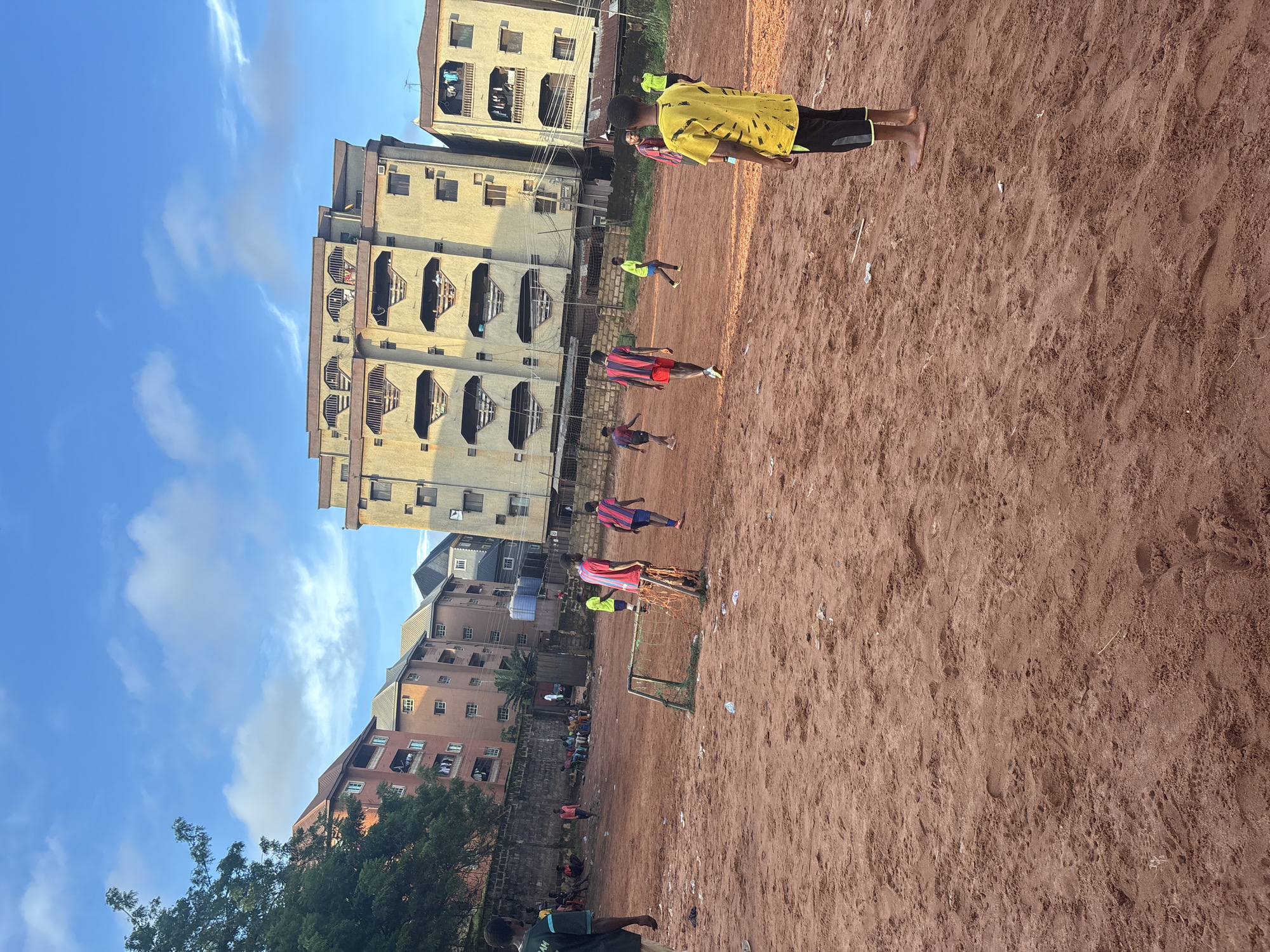 The image depicts a scene in a city, specifically identified as Onitsha, Nigeria. The setting is an outdoor urban area with a reddish-brown dirt ground, likely a makeshift soccer field. Several young men are engaged in a soccer game. One individual, wearing a yellow t-shirt with black detailing and black shorts, stands prominently in the foreground, facing away from the camera. In the background, various multi-story buildings, characteristic of an urban environment, are visible. Some of these buildings appear to be residential or commercial structures. The sky above is blue with scattered clouds, suggesting daylight. The overall atmosphere is one of casual sport and urban life.