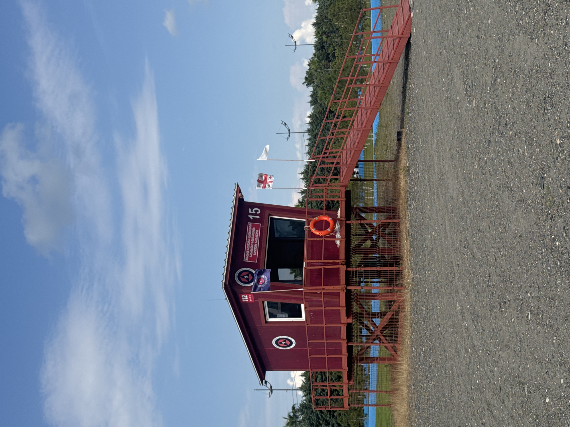 A red lifeguard station stands on a pebble beach in Anaklia, Georgia. The station is elevated on wooden stilts and accessed by a red metal staircase. Two flags fly from the station: a dark blue flag with a circular emblem and a white flag. A life preserver ring is attached to the side of the station. Trees form a backdrop behind the station, and a clear blue sky with scattered clouds is visible above.