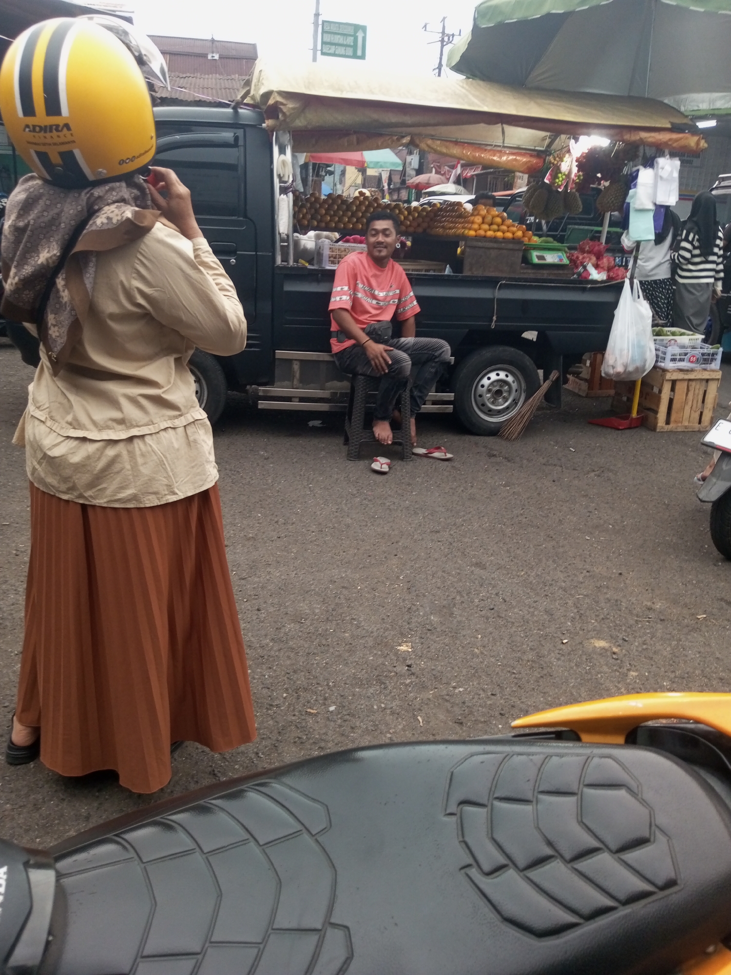 A woman wearing a yellow helmet and a tan abaya and brown skirt stands in front of a black truck. The truck is loaded with fruit, including oranges and durian. A man in a pink shirt and dark pants sits on a stool next to the truck. The scene appears to be a street market.