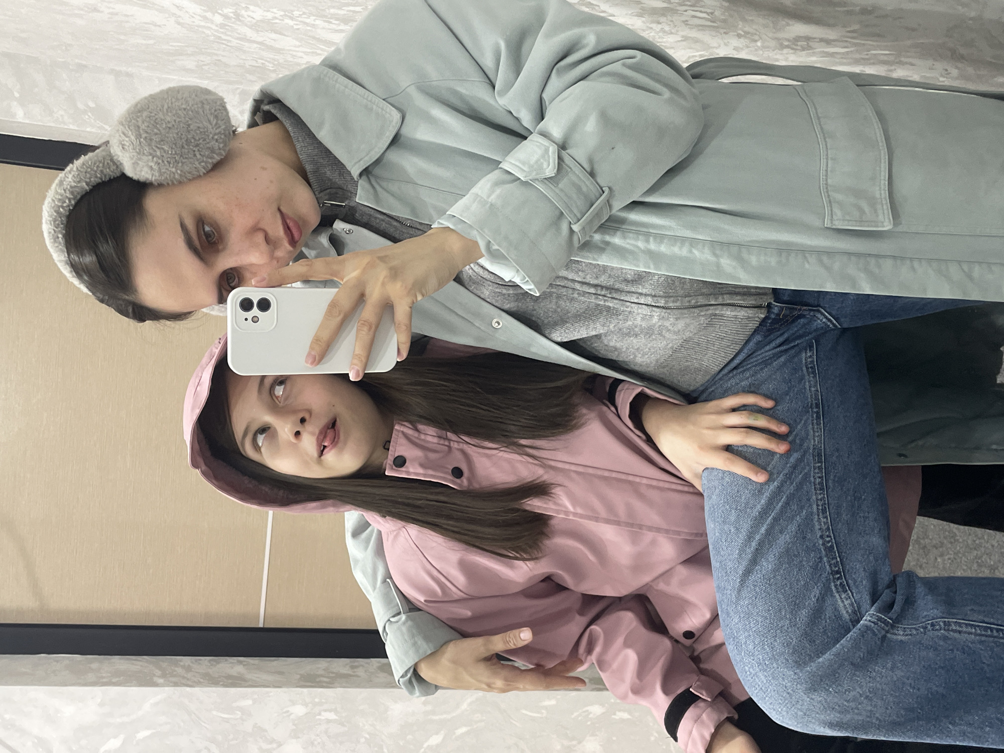 An indoor mirror selfie, taken in Izhevsk, Russia, depicts two females. The individual on the right, an adult with dark hair styled away from her face, wears grey fuzzy earmuffs, a grey turtleneck sweater, a light blue long coat, and blue jeans. She holds a white smartphone with her right hand, angled to capture the reflection, and her left arm rests around the shoulder of the other individual. The individual on the left, an adolescent with long dark hair, wears a pink hooded jacket and is seen with crossed eyes and her tongue partially extended. The background consists of a textured light grey wall on the left and a plain light beige panel on the right, separated by a dark vertical frame.
