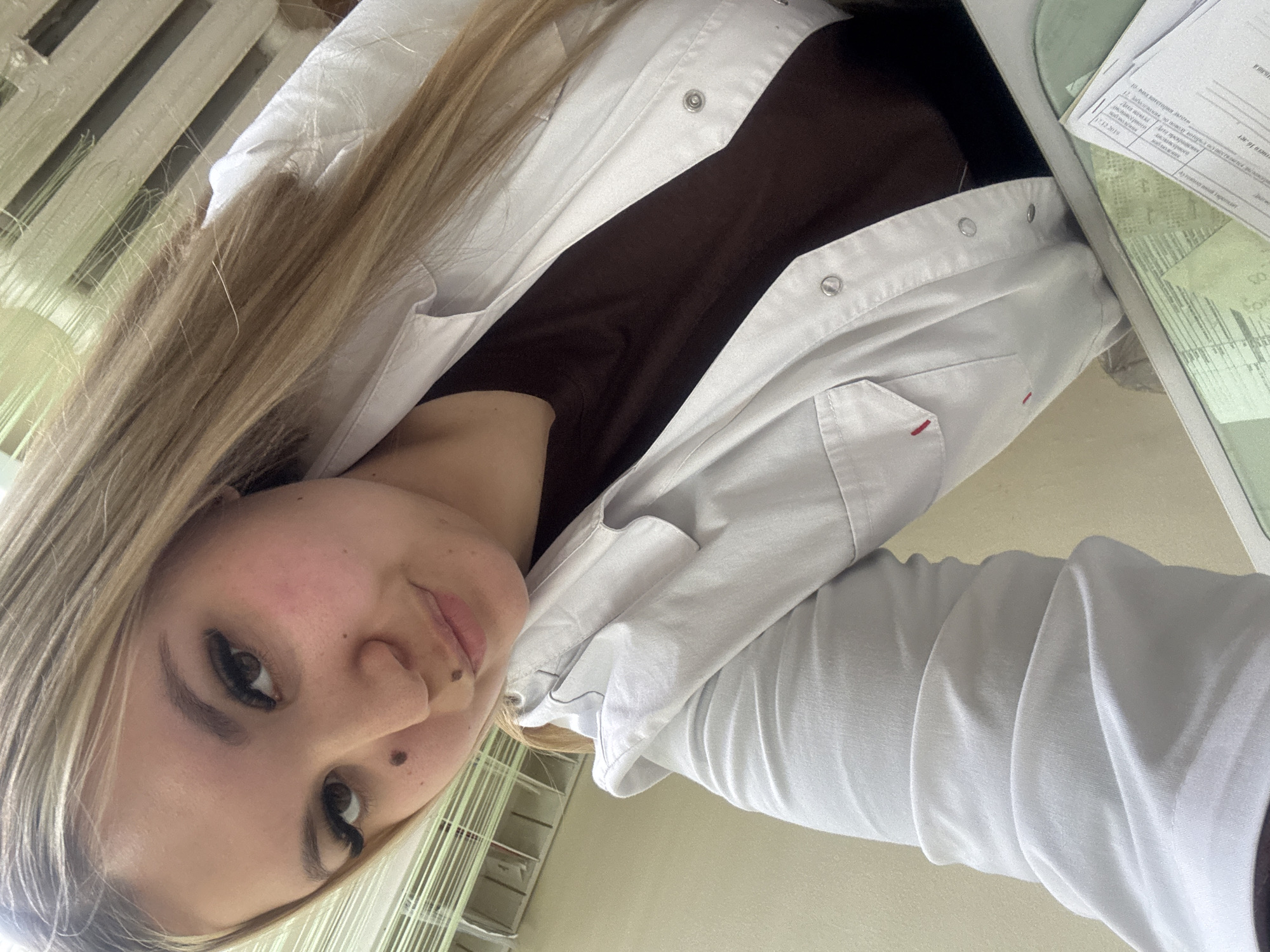 A woman with long blonde hair, wearing a white button-down coat over a dark top, is looking at the camera. She has dark eyelashes and several moles on her face. To her right, a document lies on a table. The location cannot be determined from the image.