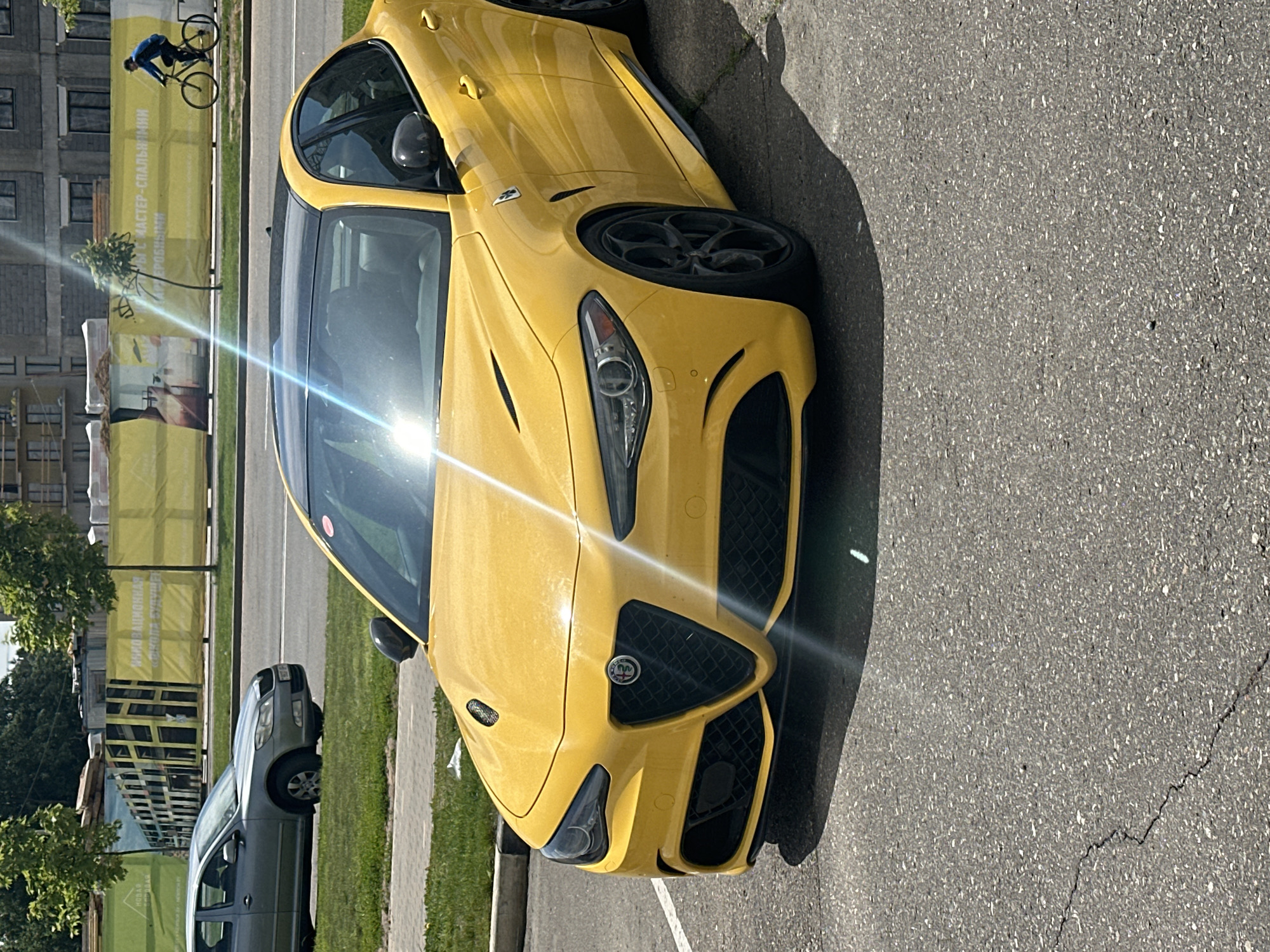The image depicts an outdoor urban scene in Бараўлянскі сельскі Савет, Belarus, under bright sunlight.
In the immediate foreground, a bright yellow Alfa Romeo Giulia Quadrifoglio sedan is parked diagonally on asphalt, facing towards the viewer's left. The vehicle features a black roof, black multi-spoke wheels, black side mirrors, and black aerodynamic elements including front grille inserts and side skirts. The circular Alfa Romeo emblem with a red Quadrifoglio cloverleaf is visible on the center of the distinctive V-shaped front grille. The asphalt surface exhibits minor cracking and is composed of variegated aggregate. A strong lens flare originating from the upper center of the image casts a bright streak across the vehicle's hood and windshield.
In the midground, to the left of the yellow Alfa Romeo, a strip of green grass and a concrete curb separate the parking area from a road. A grey second-generation Skoda Octavia hatchback is partially visible, positioned on the far side of the grass strip, facing left. Its front section and left wheel are discernible.
The background reveals a construction site. A paved road runs horizontally across the mid-background. Behind this road, a long construction fence or barrier is adorned with large banners. The banners are green on the left and yellow on the right. Cyrillic text is visible on these banners; notable phrases include "НОВАЯ БАРАВАЯ" (New Baravaya) on the green section and phrases pertaining to "МАСТЕР-СПАЛЬНИМИ" (master-bedrooms) and "ОНОВАЦИОННАЯ ШКОЛА" (innovative school) on the yellow sections. The banners also display graphic renditions of modern building facades. Beyond the fencing, incomplete concrete structures of buildings under construction are visible. A person wearing a blue top and dark pants is cycling a road-style bicycle on a paved path parallel to the road, moving from left to right on the far right side of the background.
The scene is brightly illuminated, with the sun's position creating prominent reflections and a visible lens flare, indicating clear weather.