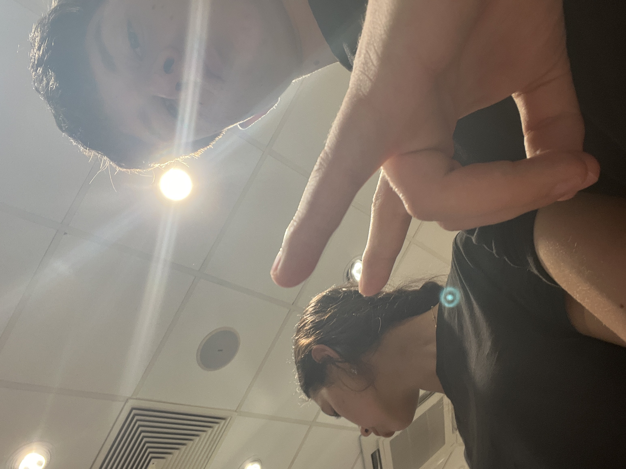 The image shows two individuals in an indoor setting, presumably an office or commercial space, indicated by the ceiling tiles and ventilation system visible in the background. One person, with dark hair, is positioned above the other, creating an over-the-shoulder perspective. A hand is also present in the frame. The other person, a woman with dark hair pulled back in a ponytail, is looking down. The location is not Popovitsa, Bulgaria, as there's nothing within the image which indicates its location.