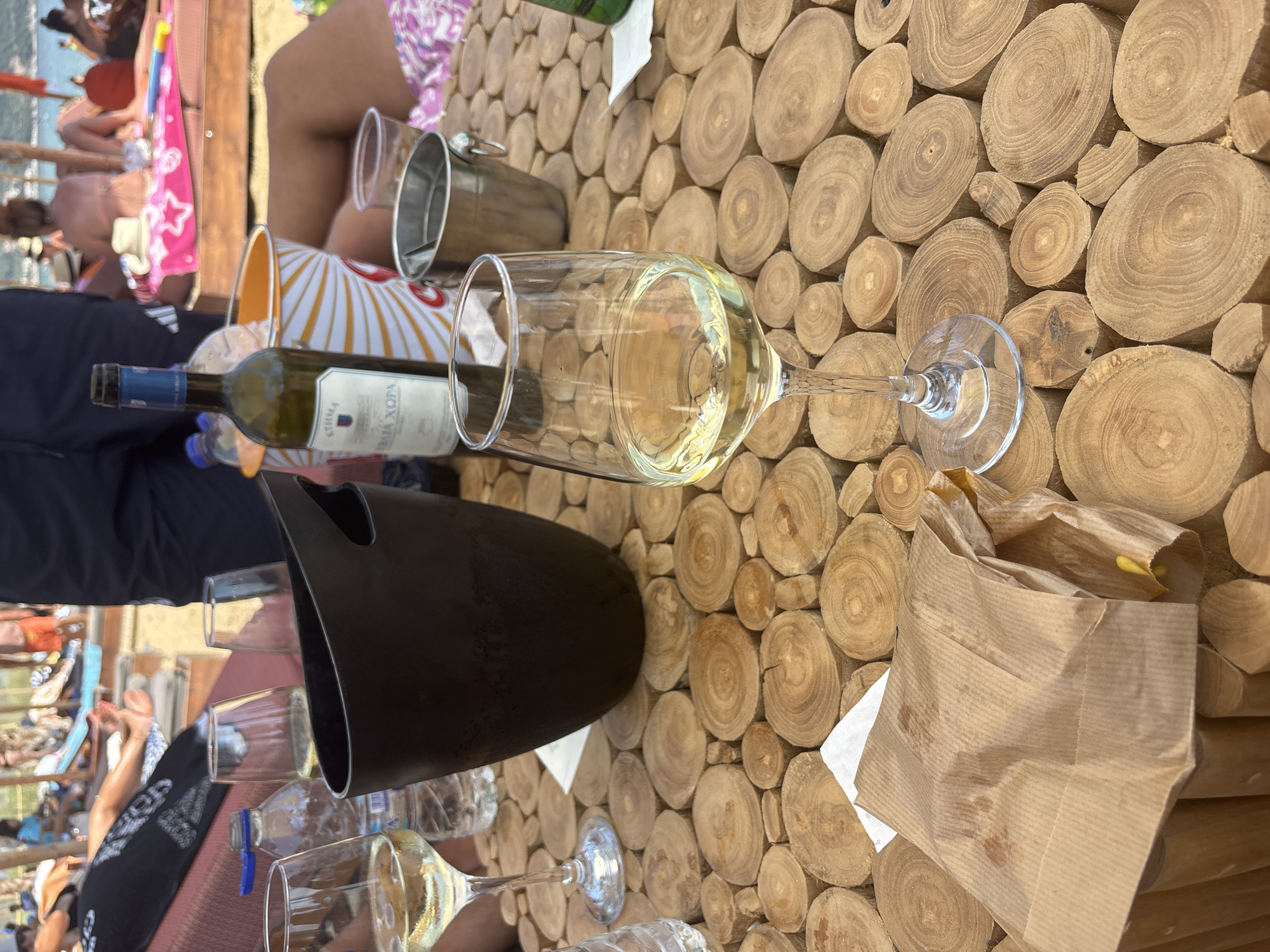 The image depicts a close-up view of a wooden table at a beachside establishment in Nea Iraklitsa, Greece. On the table, there are two glasses of white wine, one of which is prominently featured in the foreground, filled with pale yellow wine. A brown paper bag, possibly containing food, is placed on the left side of the table, with a small yellow object visible inside. A black ice bucket, an orange and white striped cooler bag, and a silver bucket are also present. A partially visible bottle of white wine with a blue label stands behind the cooler bag. In the background, several people are seen relaxing on lounge chairs on the beach, some partially obscured by the table and other objects. The environment suggests a casual, sunny day at the seaside.