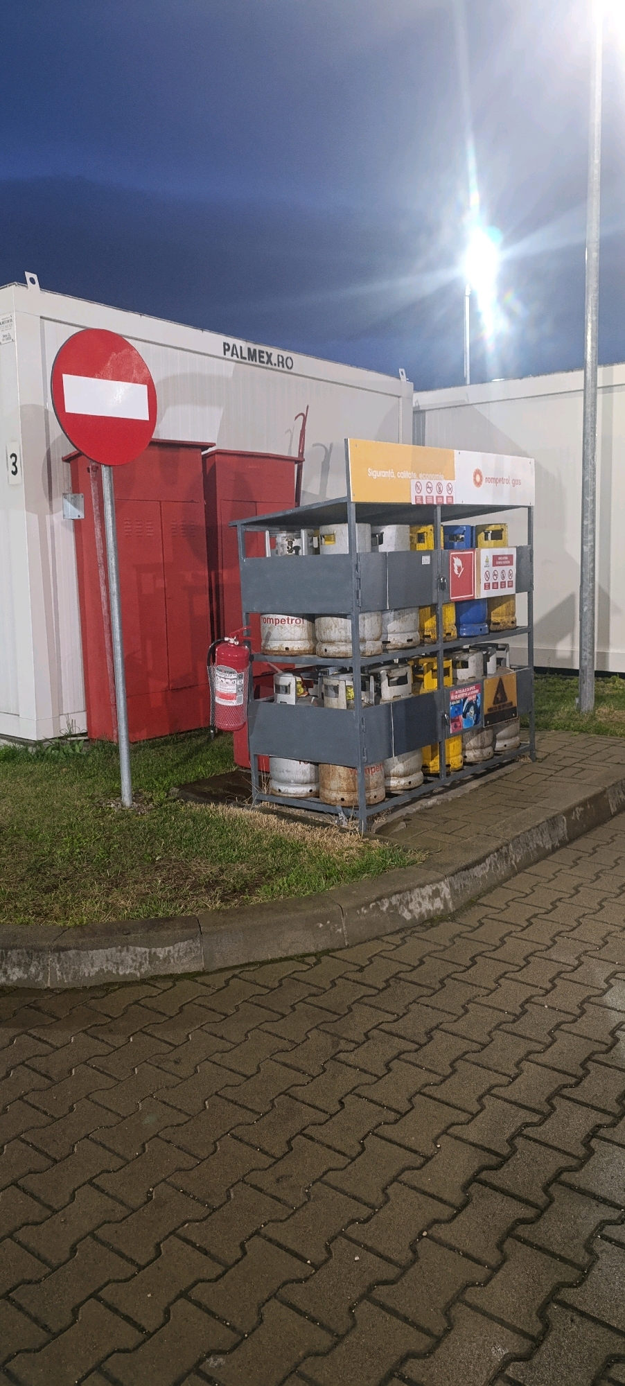 The image captures an outdoor scene at what appears to be a fuel station or a storage area, likely in Câmpulung Moldovenesc, Romania, given the context. The time of day is dusk or night, with artificial lighting casting a strong glare from the upper right.
Dominating the scene is a gray metal shelving unit filled with propane gas cylinders. These cylinders are of various sizes and colors, including white, yellow, and blue, with some showing signs of rust. The shelving unit has several tiers, and the cylinders are neatly arranged, some upright and others lying on their sides. A sign at the top of the unit reads "rompetrol gas" and features smaller icons indicating safety or service information. Below the shelving unit, to the left, is a red fire extinguisher mounted on a stand, with a mesh cage around its base.
Behind the gas cylinders and fire extinguisher, there are two tall, red metal cabinets or storage units. To the far left, a white modular building or container is visible, with the text "PALMEX.RO" printed on its side. A large circular red traffic sign with a white horizontal bar, indicating "no entry" or "do not enter," is mounted on a pole in front of this building. A number "3" is visible on the side of the white building.
The ground is paved with interlocking brick pavers, which appear wet, suggesting recent rain or cleaning. A raised concrete curb separates the paved area from a strip of dry, patchy grass. In the background, the sky is dark blue, indicating evening or nighttime. A bright light source, possibly a streetlamp or floodlight, is prominently positioned in the upper portion of the image, creating a significant lens flare effect. There are no people visible in the frame. The overall impression is one of functional storage and service infrastructure.