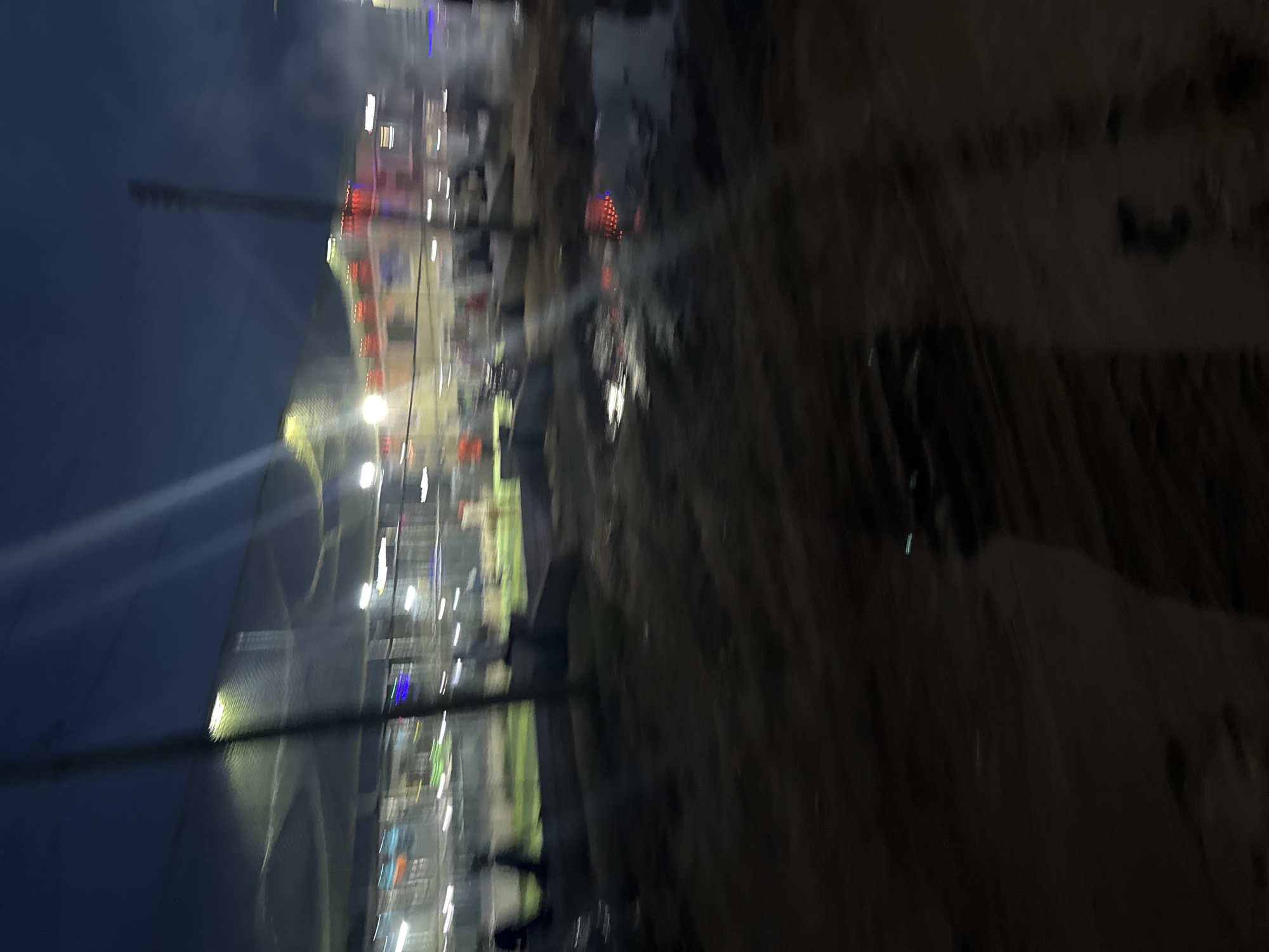 The provided image is significantly blurry and appears rotated 90 degrees clockwise. When viewed in the correct orientation, it depicts an outdoor scene at night in Ihiagwa, Nigeria.
The environment is illuminated by numerous artificial lights, creating streaks and blurs across the frame. On the left, a large, dark structure, possibly a building or tent with a curved roof, stands against the dark night sky. A tall, slender pole or mast is visible in the upper left quadrant.
The central and mid-right portions of the image are filled with bright, diffused lights, primarily white, yellow, and some red and blue. These lights suggest an active area, such as a market, fairground, or event space, with various stalls or establishments. The ground in this lit area shows hints of unevenness and possibly some vegetation.
The far right side of the image is dominated by a dark, textured surface, appearing to be rough or unpaved ground. The blur makes it difficult to discern specific objects, people, or activities. However, the presence of many lights indicates human presence and activity. The overall impression is a vibrant but indistinct nighttime gathering or commercial area. No discernible text is visible.