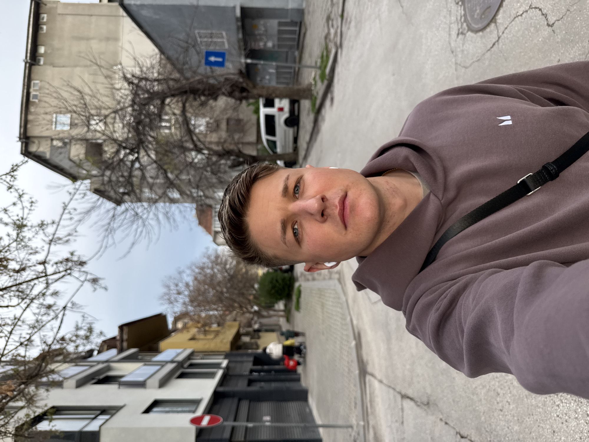 A selfie shows a man wearing a brown hoodie with a black strap across his body. He stands on a sidewalk beside a street in Plovdiv, Bulgaria. Buildings line the street, with bare trees scattered among them. A white van is parked further down the street. A street sign is visible on the side of one of the buildings. The sidewalk is gray with cracks.