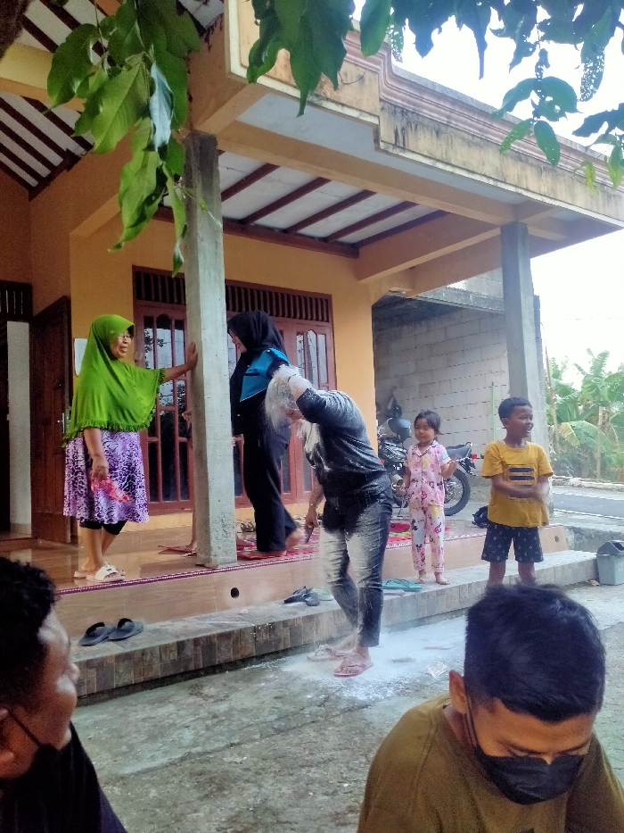 The image captures an outdoor event in Celep, Indonesia, taking place on a concrete ground area adjacent to a residential building. In the foreground, two adult males are partially visible; the male on the left is seen from the side, while the male on the right wears a black face mask and an olive-green shirt, looking downwards.
Centrally, an adult individual with short dark hair, wearing a dark long-sleeved top and dark jeans, is heavily covered in white powder and appears wet. This individual is bending forward, standing barefoot within a significant spread of white powder on the ground. Multiple pairs of footwear, including black slippers, are visible on the elevated porch steps.
On the elevated porch of the house, an adult female in a bright green hijab and a purple patterned skirt stands barefoot, smiling and leaning against a light gray concrete pillar. Behind the powdered individual, another adult female wearing a black hijab and dark attire stands barefoot on a red patterned mat on the porch.
Two children observe the scene from the ground level on the right. A young girl in pink floral pajamas stands behind the powdered individual, near a dark motorcycle parked on the unpaved area. A young boy in a yellow striped t-shirt and dark shorts stands closer to the powdered individual, smiling towards the left. A gray bucket is visible to the far right.
The residential building features a light orange facade with brown wooden windows and doors. An extended roof, supported by the central concrete pillar, covers the porch. Green leaves from an overhead tree partially obscure the upper left corner of the frame. The background reveals additional residential structures, sparse vegetation, including banana plants, and a dirt road or path. The scene is illuminated by daylight.