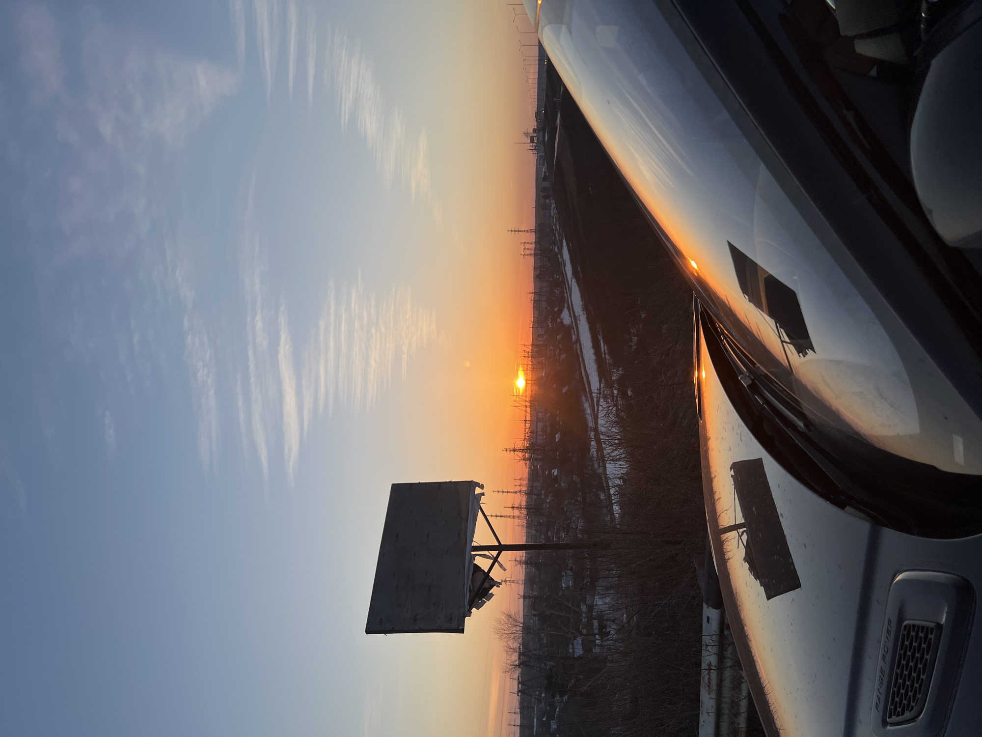 The photograph captures a sunset scene in what appears to be an industrial area near Chaikovsky, Russia. The image is oriented sideways, with the sky on the left and a vehicle's hood on the right.
Sky and Light: The sky displays a gradient, transitioning from blue at the top to orange near the horizon, where the sun is positioned. Thin clouds are visible in the blue portion.
Landscape: The landscape appears industrial, with electrical towers or pylons and possibly buildings silhouetted against the setting sun. There are barren trees or shrubs visible in the foreground.
Billboard or Sign: A dilapidated structure, possibly an old billboard or sign, is situated to the left of the sun.
Vehicle: The hood of a car is visible on the right side.
Setting: The overall setting suggests a location on the outskirts of Chaikovsky, near an industrial or utility area.