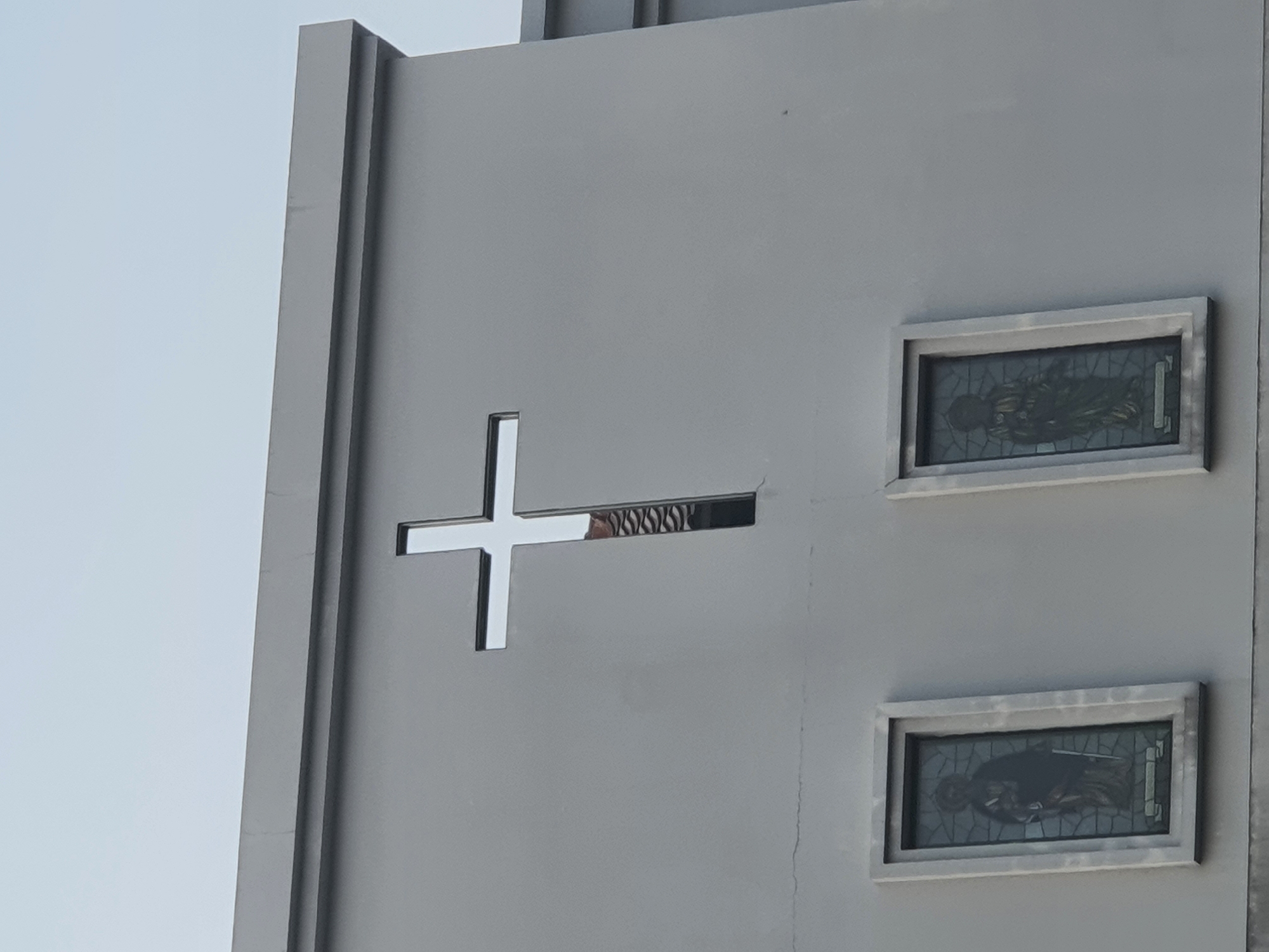 The image shows the facade of a gray building, likely a church, with a prominent cross-shaped opening near the top. Below the cross, there are two rectangular stained-glass windows, each depicting a standing figure. The sky is overcast and pale. The building is located in West Jakarta, Indonesia.