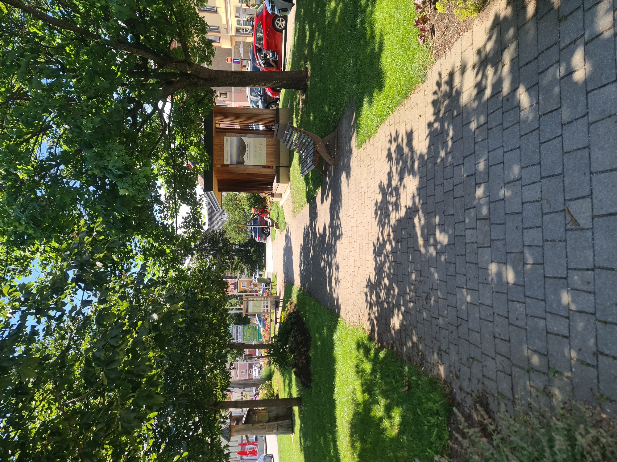 The image depicts a sun-dappled pedestrian walkway paved with rectangular gray stones, bordered by a neatly mowed green lawn on the left and right. Mature trees with lush green canopies arch over the walkway, casting intricate shadows. In the mid-ground, to the right of the walkway, is a small, modern wooden structure with a slanted roof, possibly a bus stop or information kiosk, displaying a mounted informational panel with text and imagery. Adjacent to this structure, a bench made of weathered wood slats and dark metal supports sits on the grass. Further to the left, another wooden structure, appearing older and more rustic, is partially visible. The background reveals a street with parked cars, including a bright red hatchback and a dark SUV, and buildings with pitched roofs and visible windows. Traffic signs, including a red stop sign, are also present. The overall setting suggests an outdoor public space within a town or village.