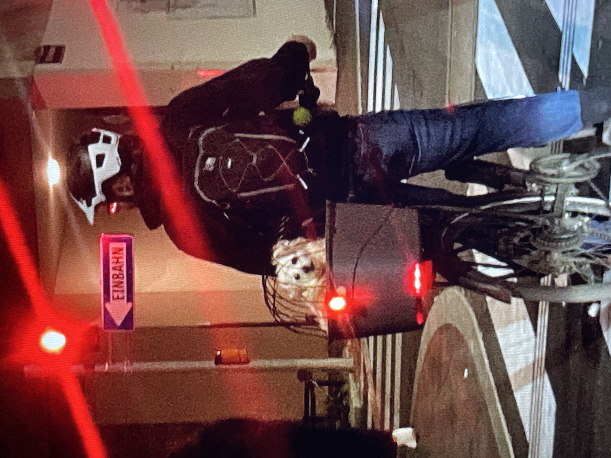 A person wearing a helmet and dark clothing is on a bicycle. The person is likely riding the bicycle on a street with a sign that reads "Einbahn" with a downward-pointing arrow. The environment is urban, as indicated by the architecture and infrastructure. The location appears to be in Germany due to the German sign.
