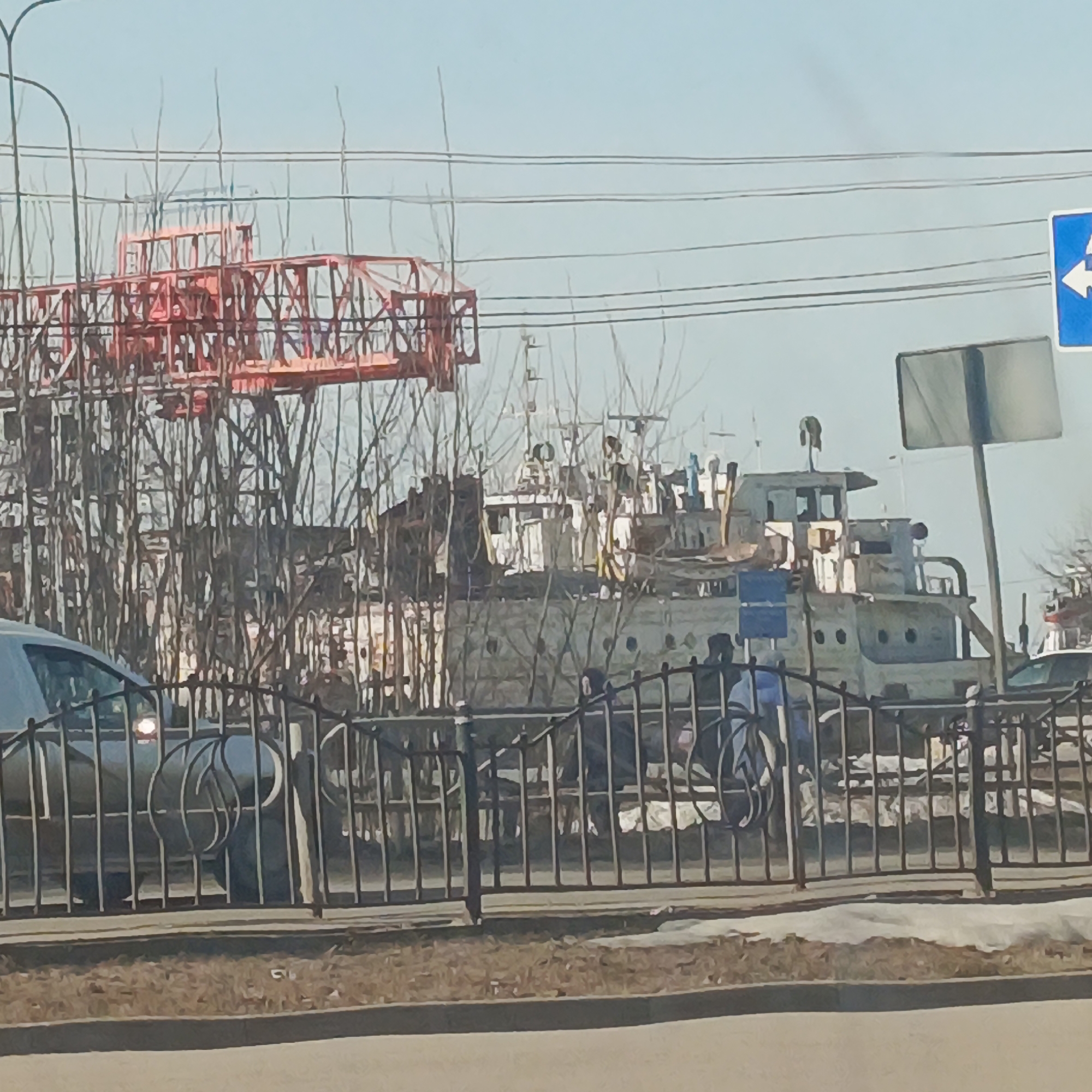The image shows a riverside scene in the city of Chaikovsky, Russia. A large, derelict river boat stands behind a black metal fence on what appears to be a riverbank. The boat is multi-tiered and predominantly white, with visible cabin windows and other nautical features. A construction crane, painted orange, is visible to the left, partially obscured by leafless trees. A signpost, and road sign (blue with a white arrow) are also visible in the scene. In front of the fence, a car is parked, with one of its headlights reflecting. Two people on bikes are also visible behind the fence. The foreground consists of a paved road and a patch of brown grass. The sky is a light blue.