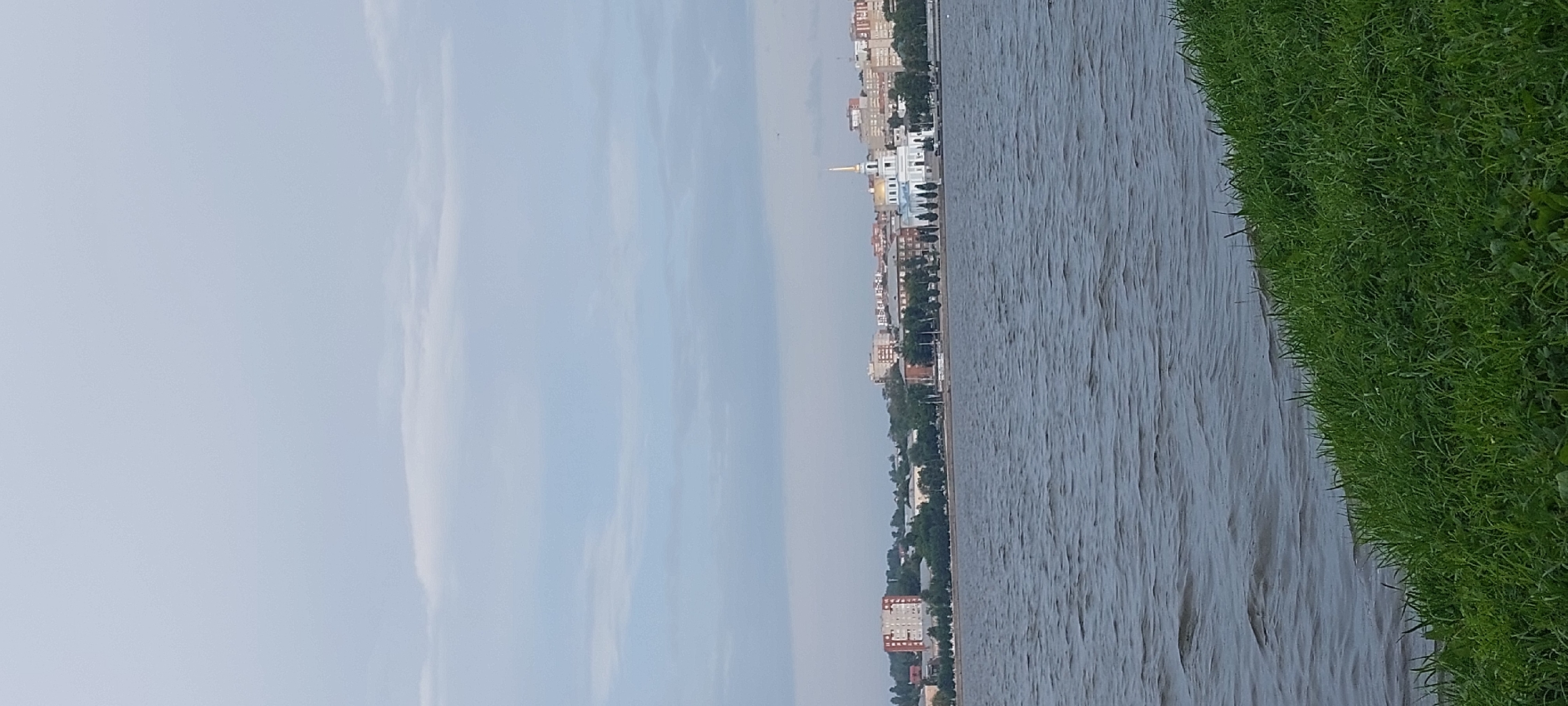 The image depicts a wide, choppy body of water in the foreground, likely a river or lake, with a prominent grassy embankment on the right side. In the background, a cityscape unfolds along the far bank of the water. The city features a mix of residential buildings and a prominent white structure with a golden dome and steeple, which is likely a church or cathedral. The sky above is overcast with a pale blue hue. The scene is a panoramic view of the Votkinsk city embankment.