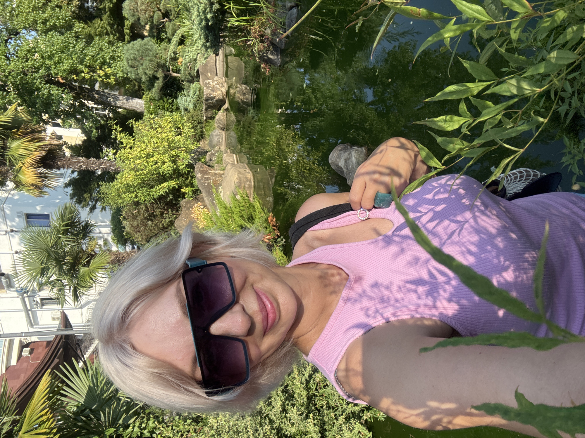 A blonde woman with short hair and large sunglasses is taking a selfie in a garden with a pond. She is wearing a pink sleeveless top. The garden features palm trees, lush green bushes, rocks, and a body of green water reflecting the surrounding foliage and a building. The scene is bathed in sunlight, creating dappled shadows on the woman and the vegetation. Based on the presence of palm trees and the style of architecture, this scene is likely located in a resort area in Sochi, Russia.