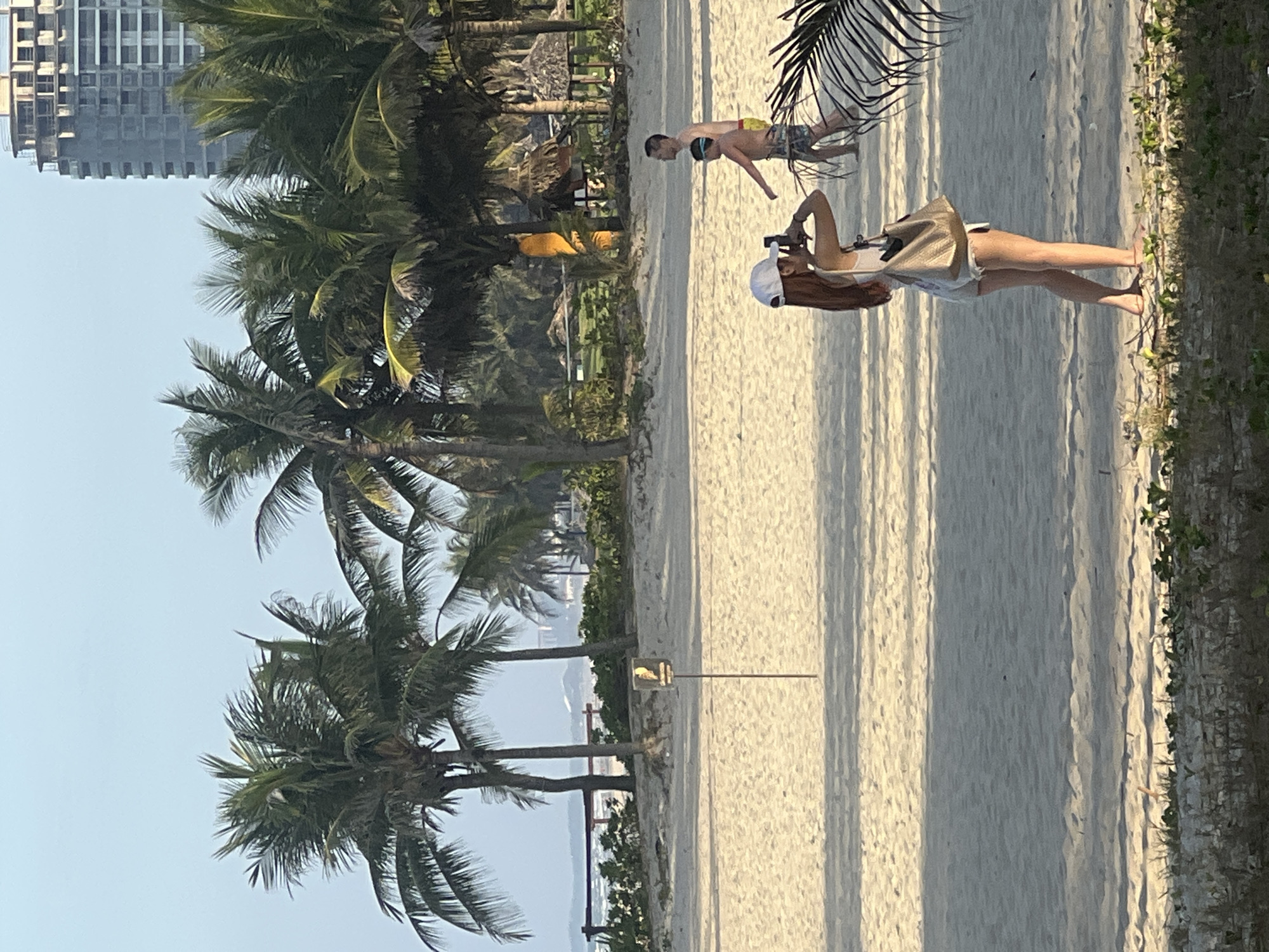 The image depicts a scene at a beach in Da Nang, Vietnam. In the foreground, a woman wearing a white baseball cap and a light-colored tank top is standing on the sand, holding a phone and appearing to take a picture. She is carrying a tan shoulder bag. Further in the background, two males are walking on the beach; one, wearing swim trunks, has a diving mask on his forehead, and the other, an adult, is shirtless. Behind them, a line of palm trees stretches across the sandy beach, with a resort area featuring thatched-roof structures and a tall, unfinished building visible in the distance under a clear sky. The ocean is visible in the far background.