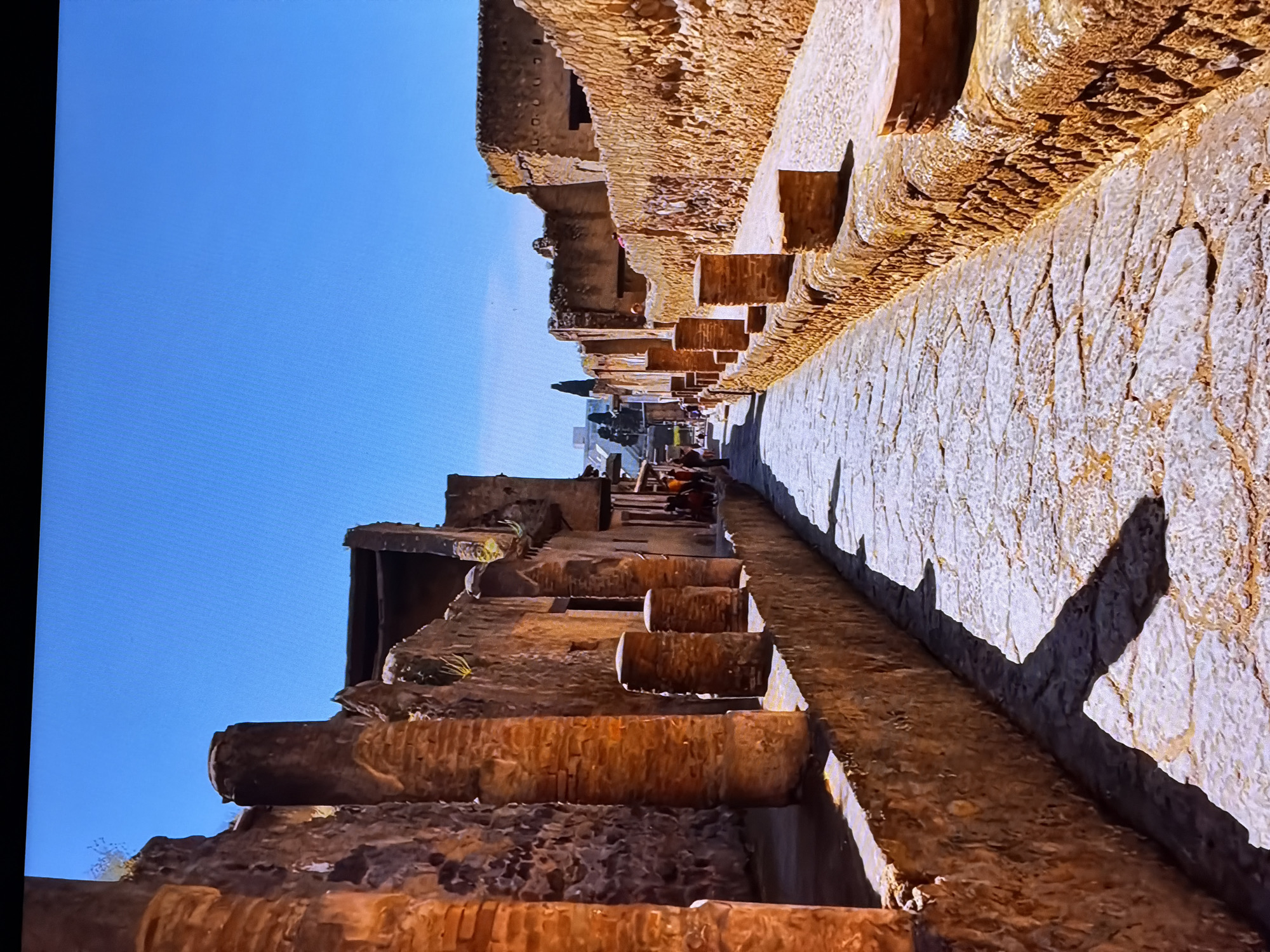 The image shows a stone-paved street lined with ancient brick structures on both sides. The structures appear to be ruins, with some columns and walls remaining. Sunlight casts long shadows across the street. There are a few people walking in the distance on the left side of the street. The sky is clear and blue. The location is identified as the city of Chirchik, Uzbekistan, which is known for its historical sites and archaeological remains.