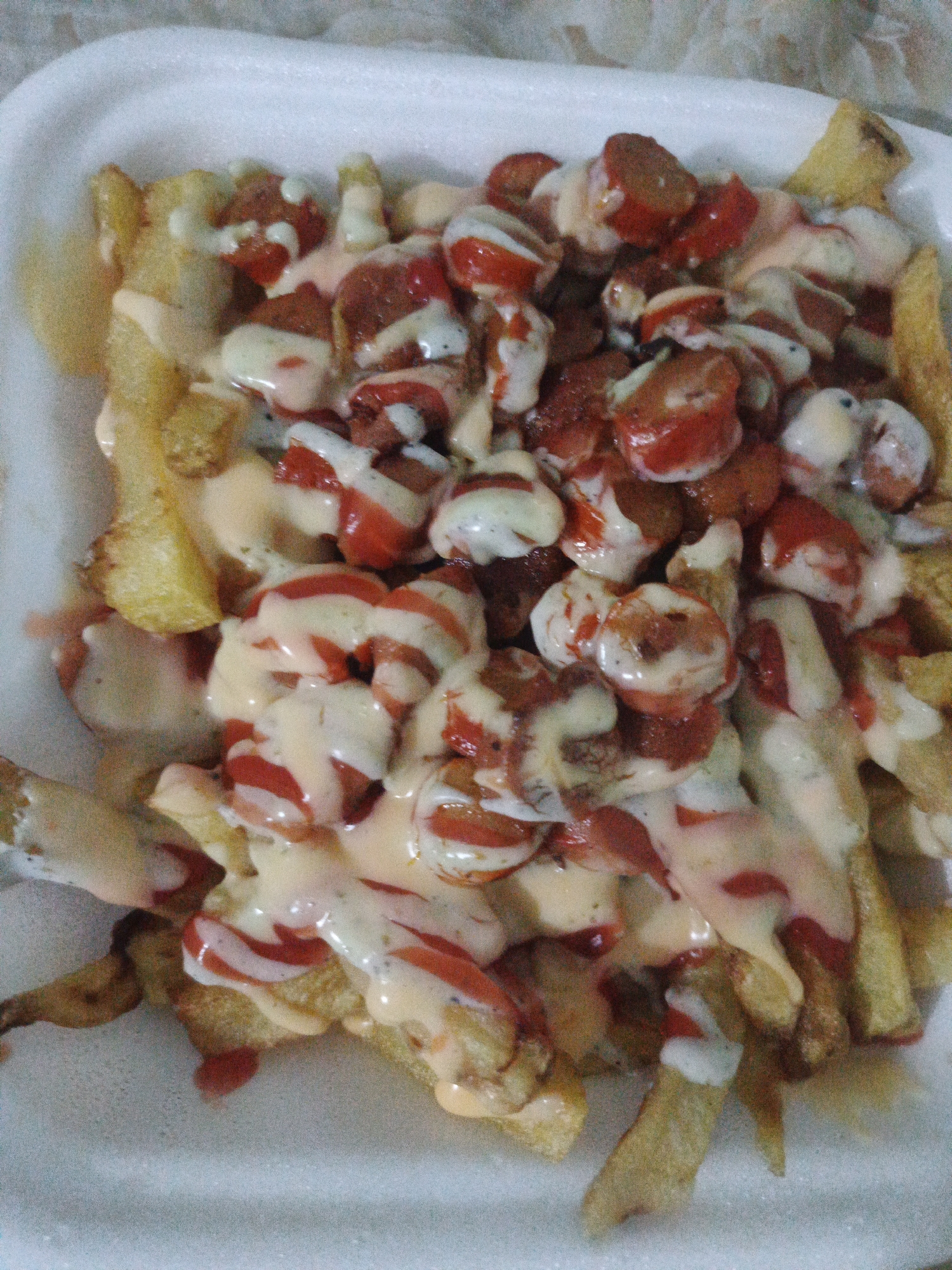The image depicts a serving of Salchipapas in a white styrofoam container. The dish consists of french fries topped with sliced hot dogs, ketchup, and mayonnaise-based sauces. The food is located in Tinaquillo, Venezuela.