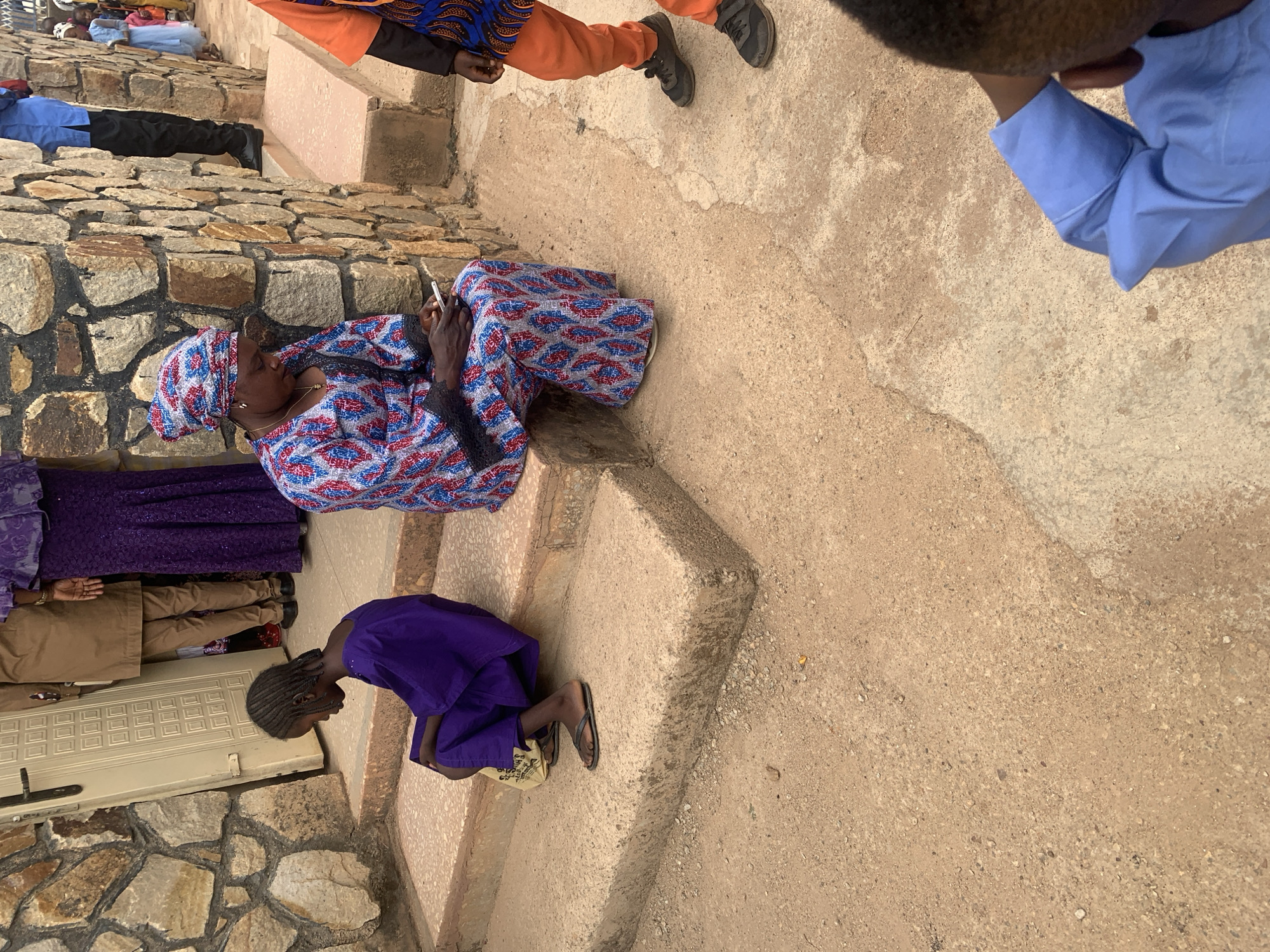 A group of people are situated near a stone structure in what is presumably the city of Jos, Nigeria. A woman is sitting on a step using a mobile phone. A young girl in a purple dress is kneeling below her. Additional people are visible in the background. The ground is composed of stone and dirt. A man wearing a blue shirt is also visible in the frame.