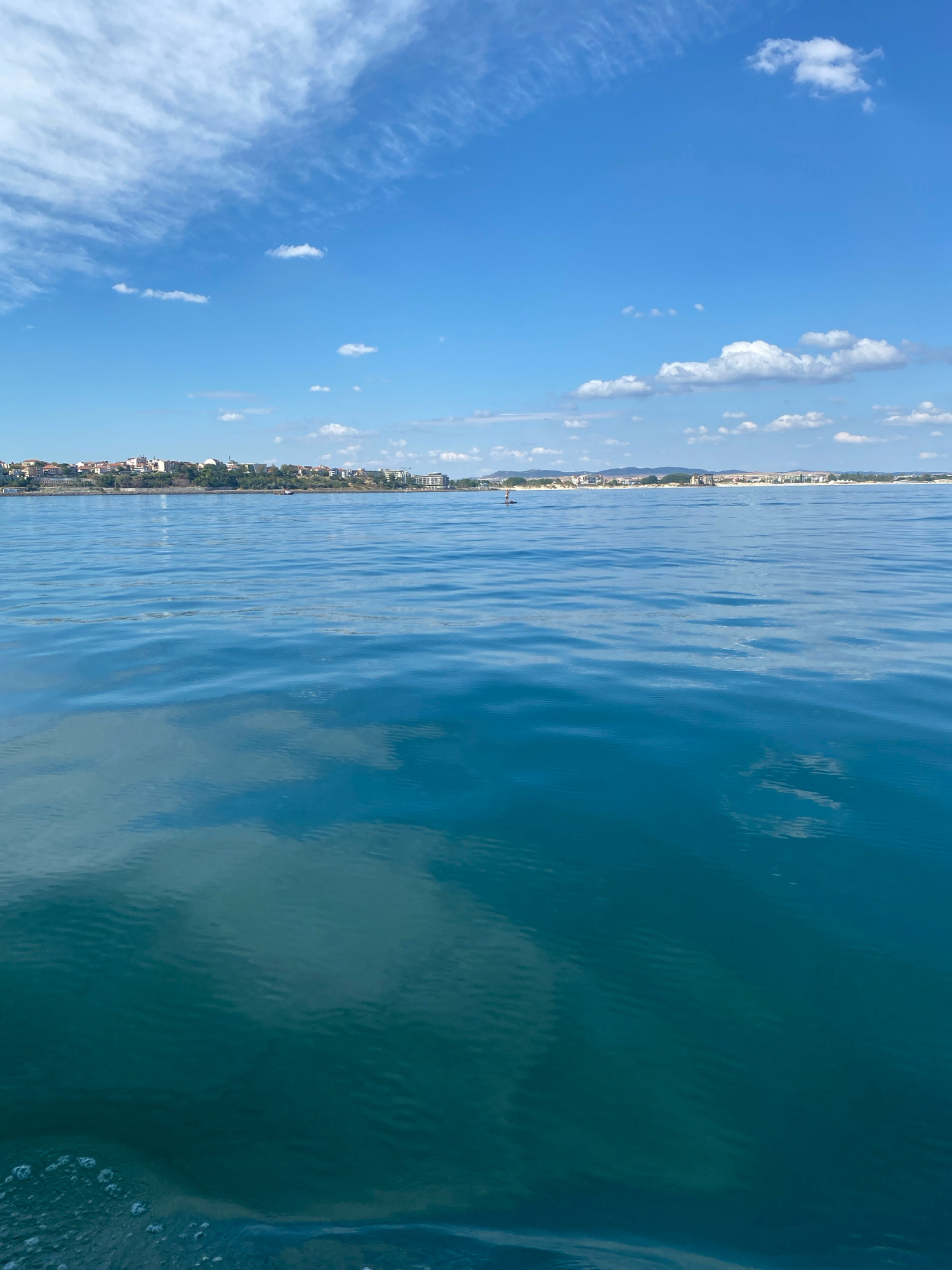 The image captures a view of the sea with the historic town of Nesebar, Bulgaria, visible in the distance along the coastline. The foreground is dominated by the calm, blue-green waters of the sea, reflecting the sky and clouds. Gentle ripples disturb the surface, and a few bubbles are visible near the bottom left corner, suggesting movement from a boat.
The middle ground features the shoreline of Nesebar, characterized by a cluster of buildings with terracotta roofs and white facades, interspersed with greenery. Some larger, modern structures are also present, indicating a mix of historical and contemporary architecture. The coastline stretches horizontally across the frame, with hills or mountains forming a backdrop further inland.
The sky above is a vibrant blue, populated with scattered white, wispy clouds, some of which are elongated and streaky, while others are more defined and puffy. The overall impression is a tranquil, sunlit day at the sea, with a prominent historical Bulgarian town as the focal point on the horizon. A solitary figure on a paddleboard is visible in the water towards the left of the frame, providing a sense of scale and human presence.