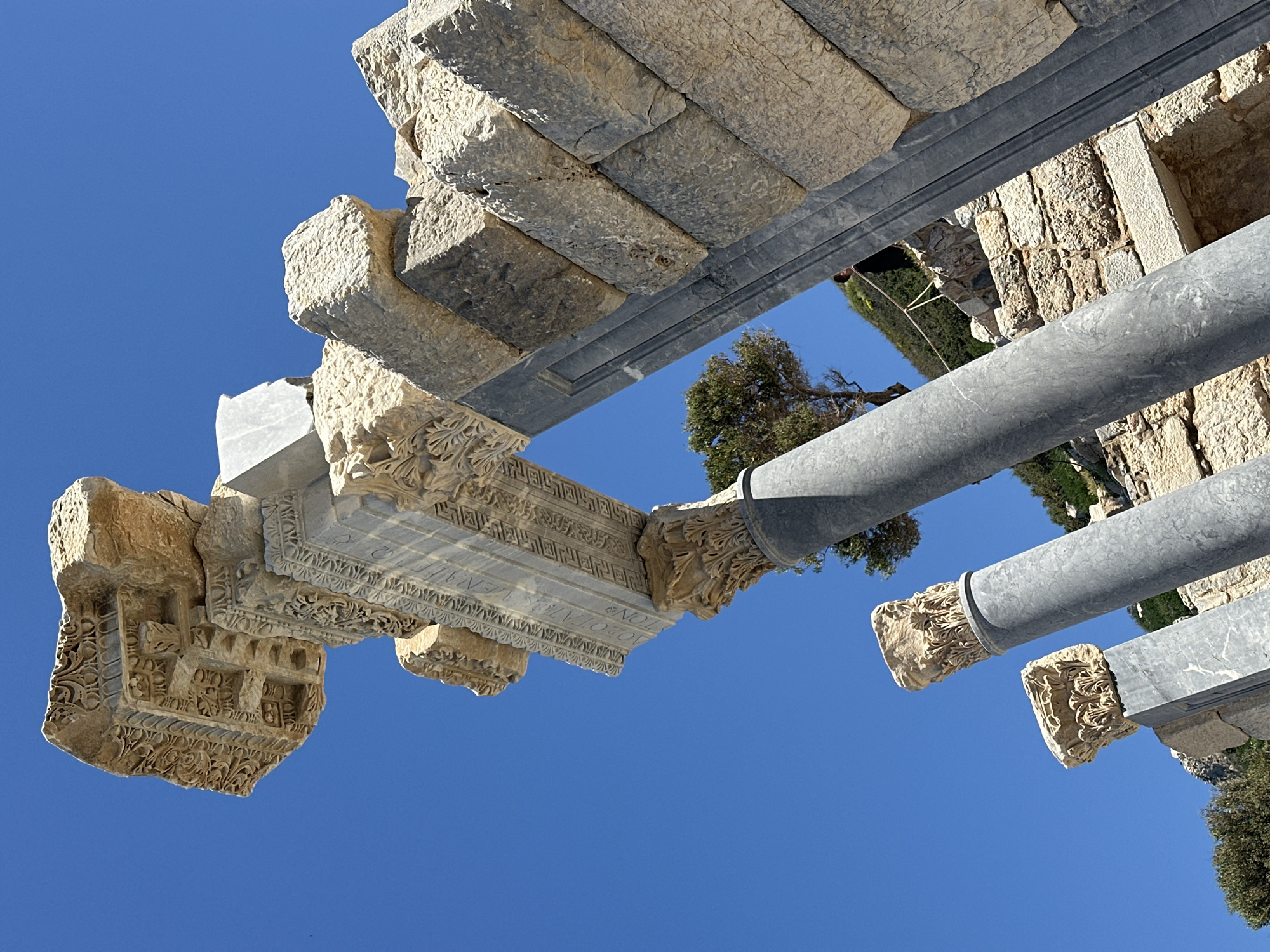 The image, taken in Datça, Turkey, presents an upward-angled view of ancient architectural ruins against a clear blue sky. The ruins consist of intricately carved stone blocks, likely remnants of a temple or similar structure. Gray cylindrical metal supports are visible, likely for modern reinforcement. Green foliage and tree branches peek through the gaps in the stone structure, adding a touch of nature to the scene. The perspective is tilted, emphasizing the height and complexity of the ruins.