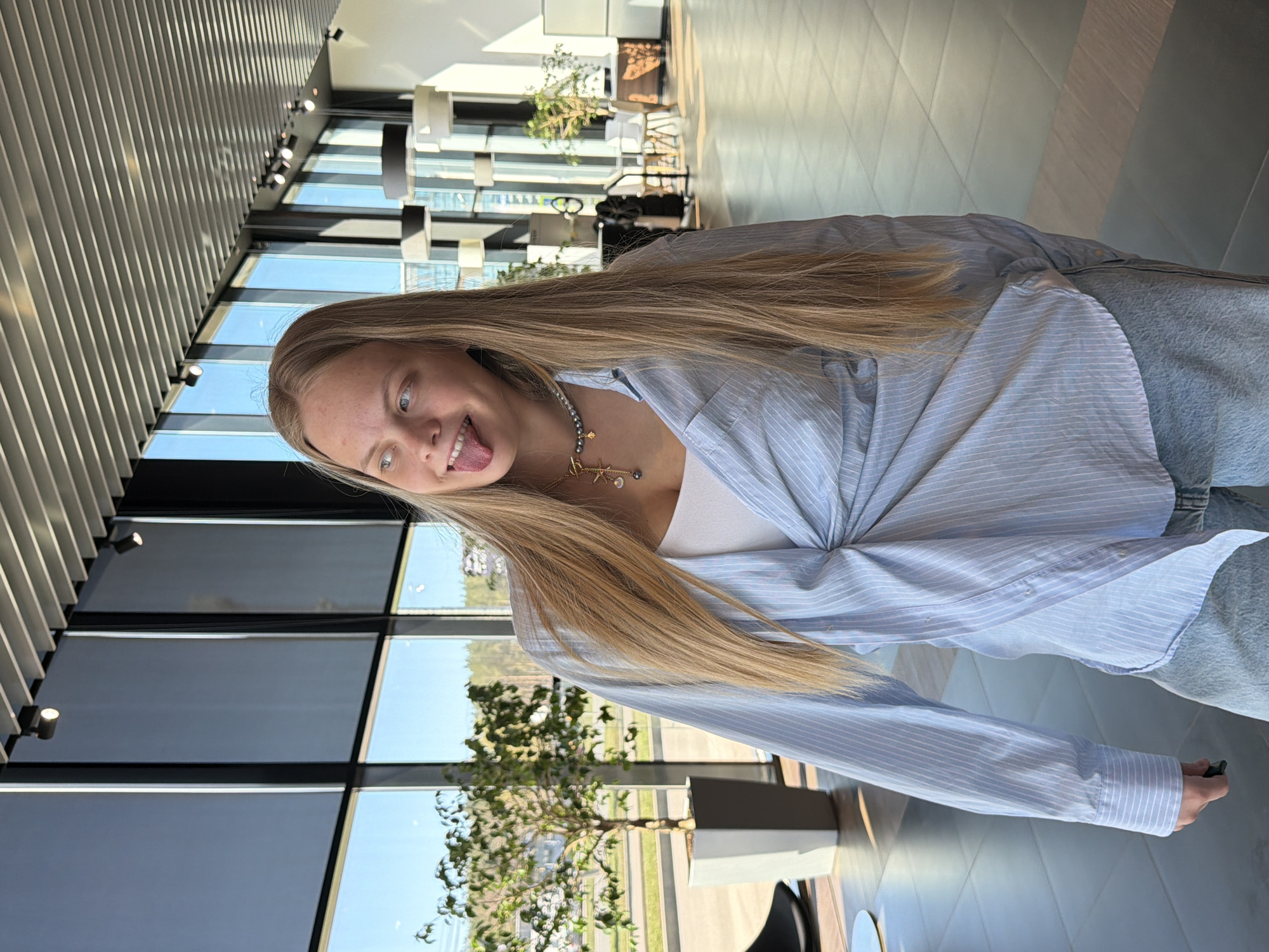 A young woman with long blonde hair sticks out her tongue in a playful manner. She is wearing a light blue striped shirt, a white tank top, and jeans. A delicate necklace adorns her neck. She is positioned indoors in what appears to be a modern building with large windows that provide ample natural light. Outside the window, greenery and possibly other buildings can be seen. The location may be Izhevsk, Russia, but this cannot be verified from the image alone.