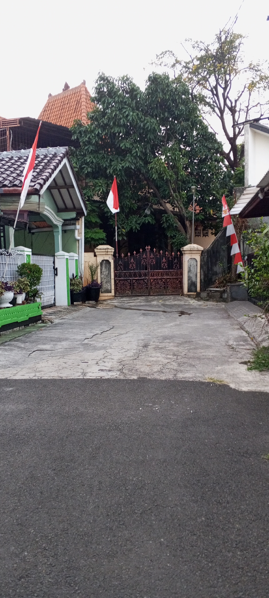 The image depicts a street scene in Depok, Indonesia, featuring a residential entrance with two gates. The foreground shows a paved asphalt road, transitioning to a cracked concrete driveway. On the left, a light green house with a tiled roof is visible, adorned with a red and white Indonesian flag. Adjacent to the house is a white fence with green accents and potted plants. The central part of the image is dominated by a dark, ornate metal gate, flanked by decorative pillars. A red and white Indonesian flag is planted on a pole to the left of the gate. To the right of the gate, another pillar stands, and a wall extends, also decorated with a red and white striped flag. Lush green trees partially obscure the background, with glimpses of houses and structures behind the foliage. The sky is overcast, suggesting a cloudy day.