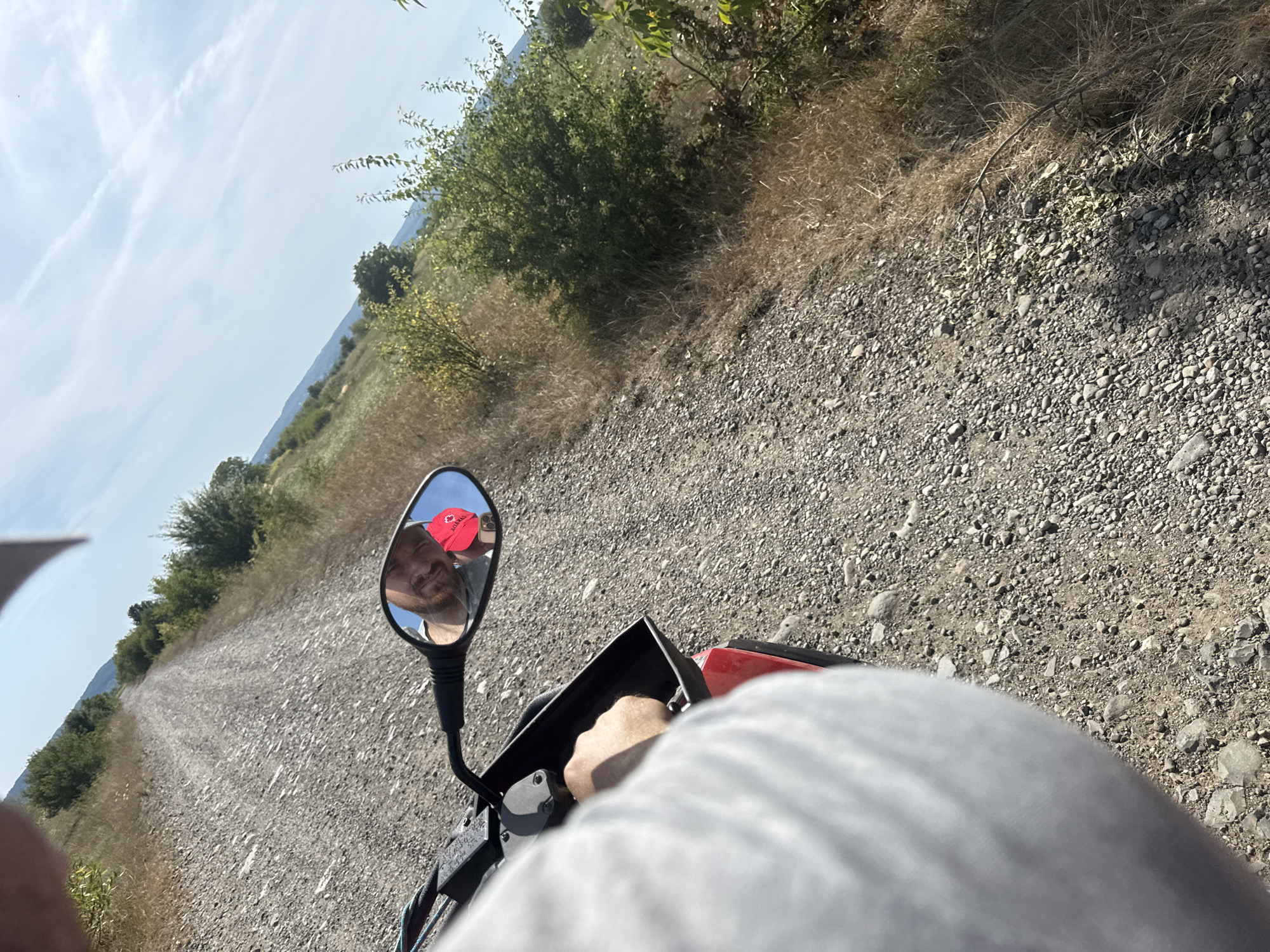 The image shows a person riding a motorcycle on a gravel road. The perspective is from the rider, looking forward. In the mirror of the motorcycle, a reflection of the rider is visible. The rider is wearing a red cap with a Canadian flag emblem. The environment appears to be rural, with dry grass and bushes on the sides of the road, and hills in the background under a clear blue sky with some wispy clouds. The date and time are not determinable from the image.