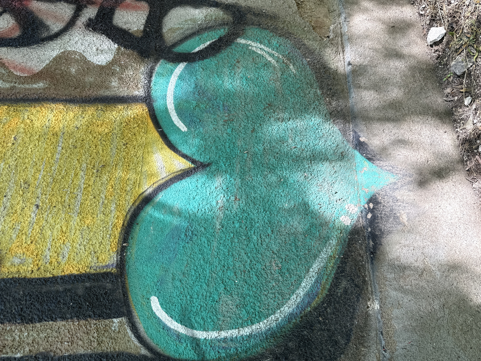 A close-up shot captures a segment of graffiti art on a concrete surface. The graffiti features a rounded, teal shape outlined in black, with white highlights adding dimension. Adjacent to the teal shape, a section is painted yellow, bordered by black lines. The concrete surface surrounding the graffiti shows signs of weathering, and debris from the environment is visible near the edge. The location of the graffiti is Pleven, Bulgaria.