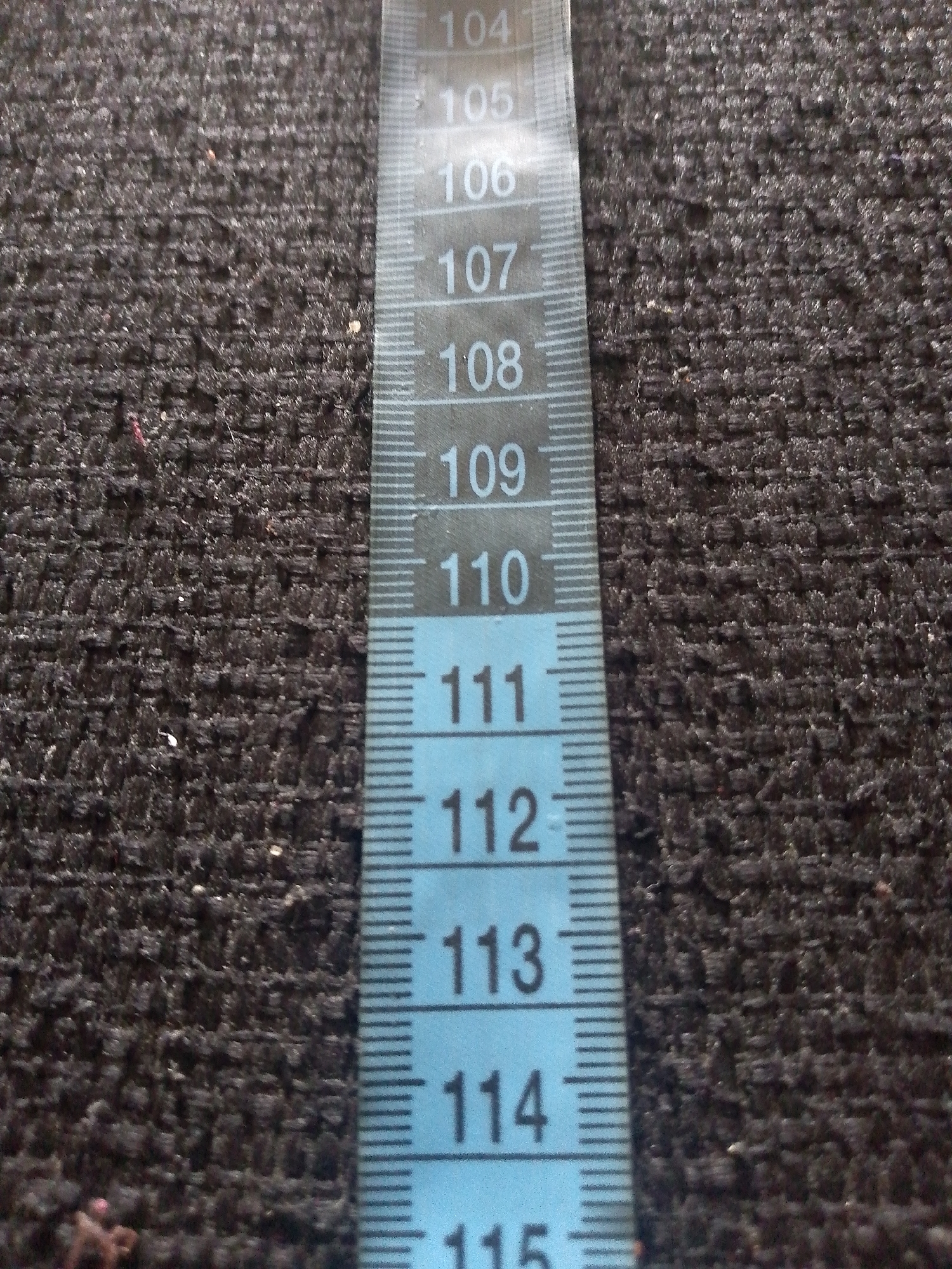 This is a close-up, high-angle shot of a blue and white measuring tape laid diagonally across a dark, textured surface, likely a fabric or carpet. The measuring tape shows markings and numbers from 104 to 115. The background is out of focus, emphasizing the detail of the measuring tape. There are no people or specific landmarks visible, and therefore no identifiable location in Jauro Yinu, Nigeria. The scene is static and appears to be an indoor setting based on the lighting.