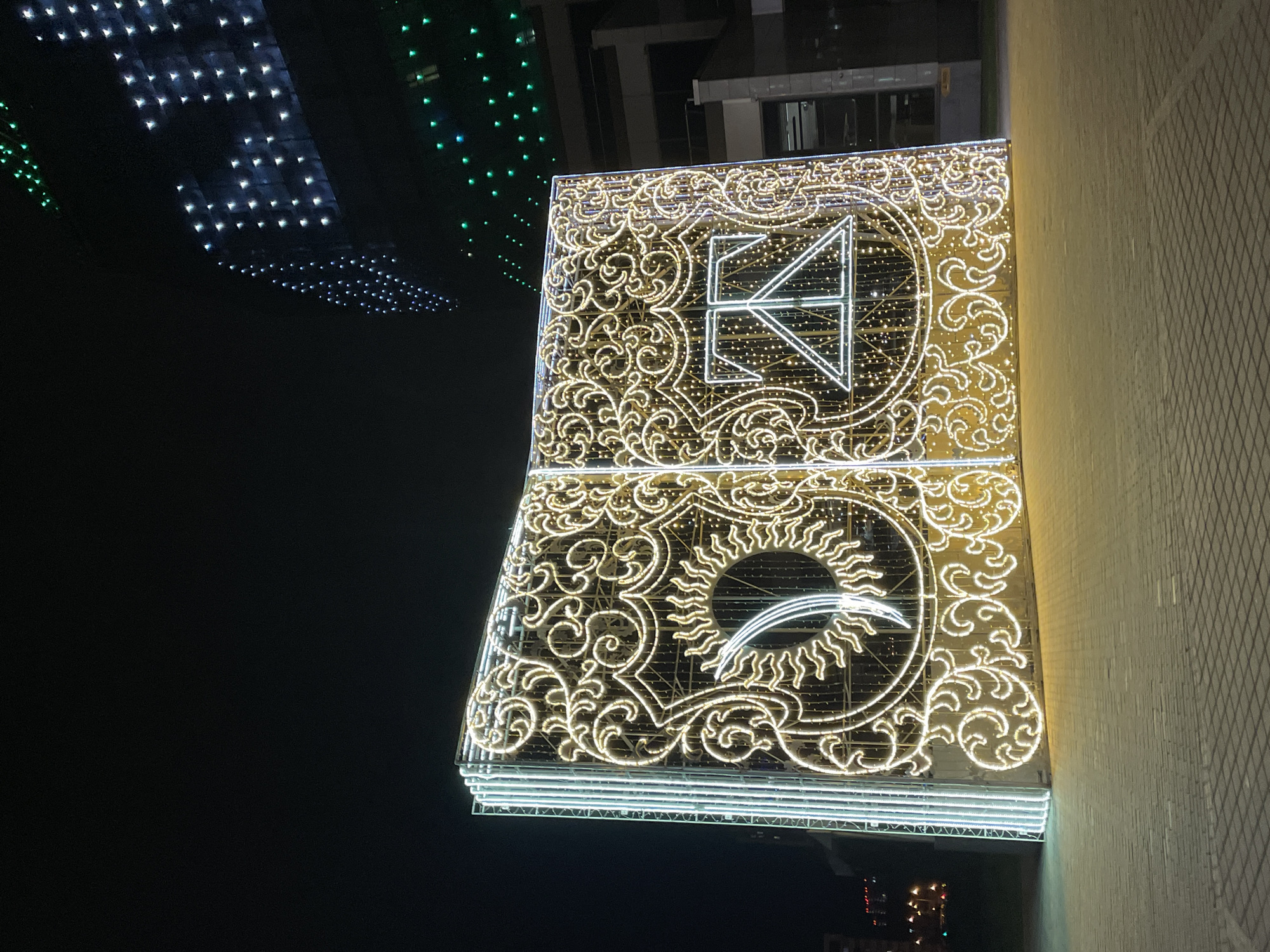 The image shows a building facade adorned with illuminated decorations in Minsk, Belarus. The decorations are gold-colored lights arranged in intricate patterns. The top section features a geometric design, while the lower section includes a sun-like image. Other buildings with decorative lights are visible in the background. The image was taken at night.