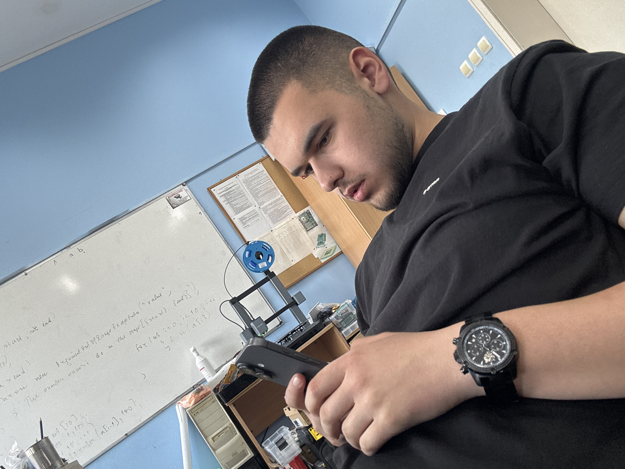 Here is a detailed description of the image:
**People:** A young man with a short haircut is the main subject. He is looking down at a smartphone he holds in his hands. He is wearing a black t-shirt and a black wristwatch.
**Objects:**
* Smartphone in the man's hands.
* A 3D printer is visible in the background.
* A whiteboard covered in hand-written text and mathematical expressions.
* An old computer tower and various electronic components.
**Environment:** The scene appears to be indoors, potentially in a classroom or laboratory. The walls are painted a light blue.
**Actions:**
* The man is looking at the smartphone.
**Interactions:**
* The man appears to be focused on his smartphone.
**Location:** It is impossible to determine the location of the scene with certainty from the image. The image does not provide any context or visual cues that would allow the location to be determined.