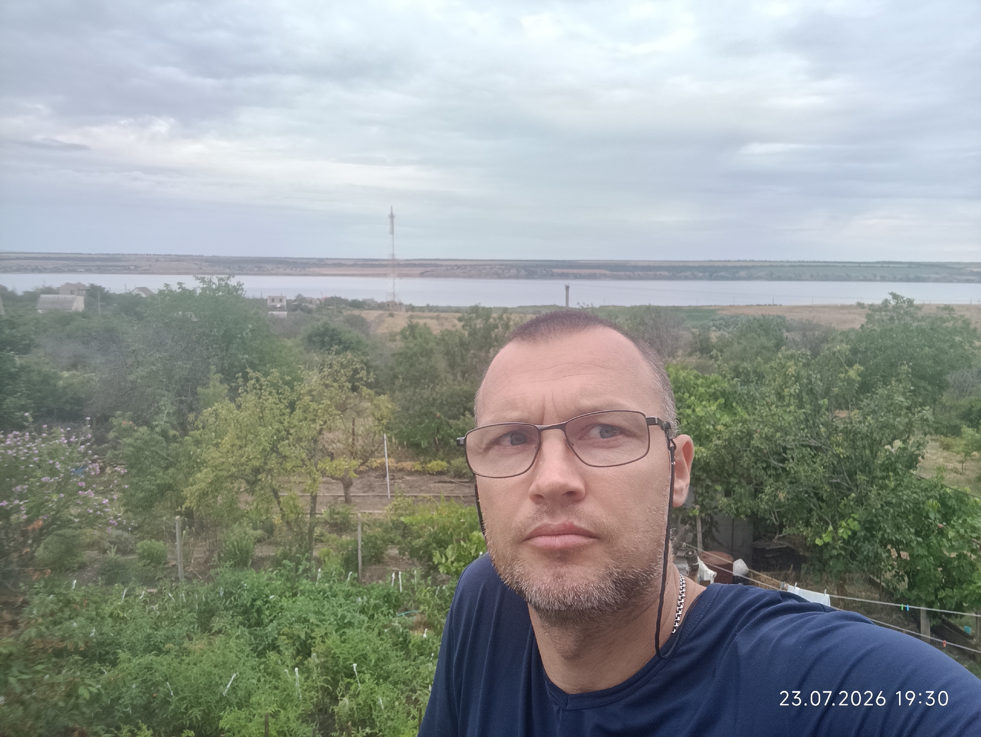 A man wearing glasses and a dark blue t-shirt is taking a selfie outdoors. He has short, graying hair and a short beard. Behind him, there is a view of a body of water and a distant landscape with a tower. The foreground is filled with lush green vegetation, including trees and bushes, suggesting a rural or garden setting. The image is dated "23.07.2026 19:30".