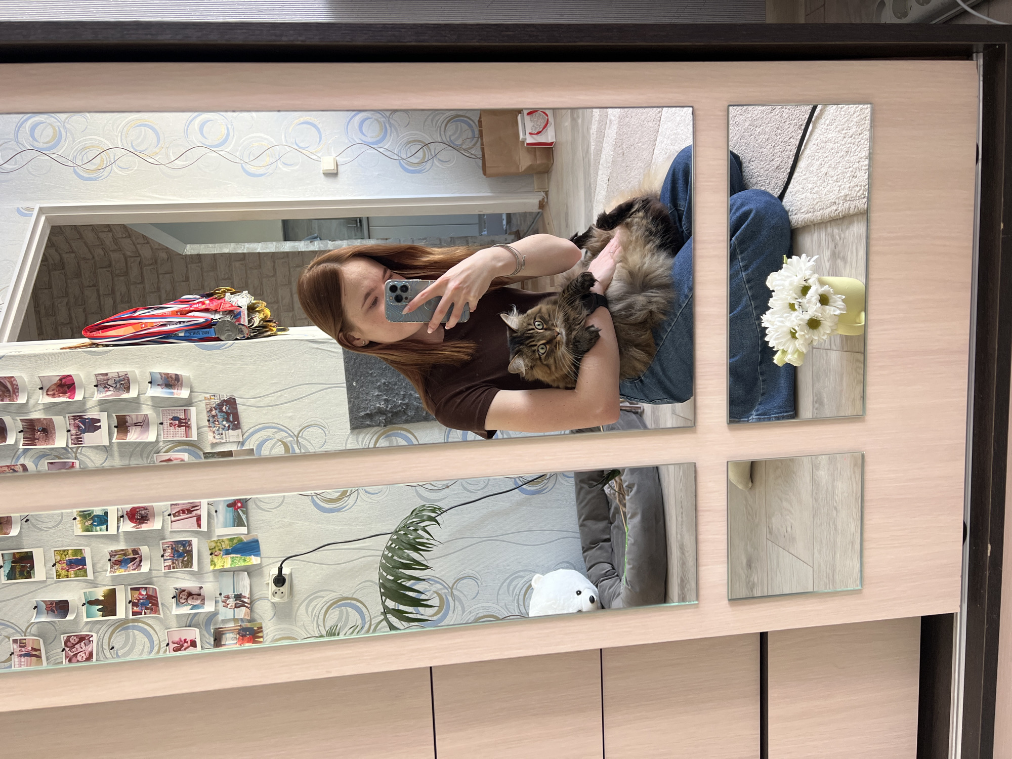 A woman is holding a cat in front of a mirror. The mirror is composed of several square and rectangular mirrors arranged together. The woman is wearing a brown shirt and blue jeans. She is taking a photo of herself and the cat with her cell phone. The cat is a brown and black Maine Coon. Behind the woman is a wall with patterned wallpaper, decorated with small photos and medals. A flower vase is visible in the top right corner. A white stuffed animal and a houseplant are visible in the bottom left corner. The location is Chaikovsky, Russia.