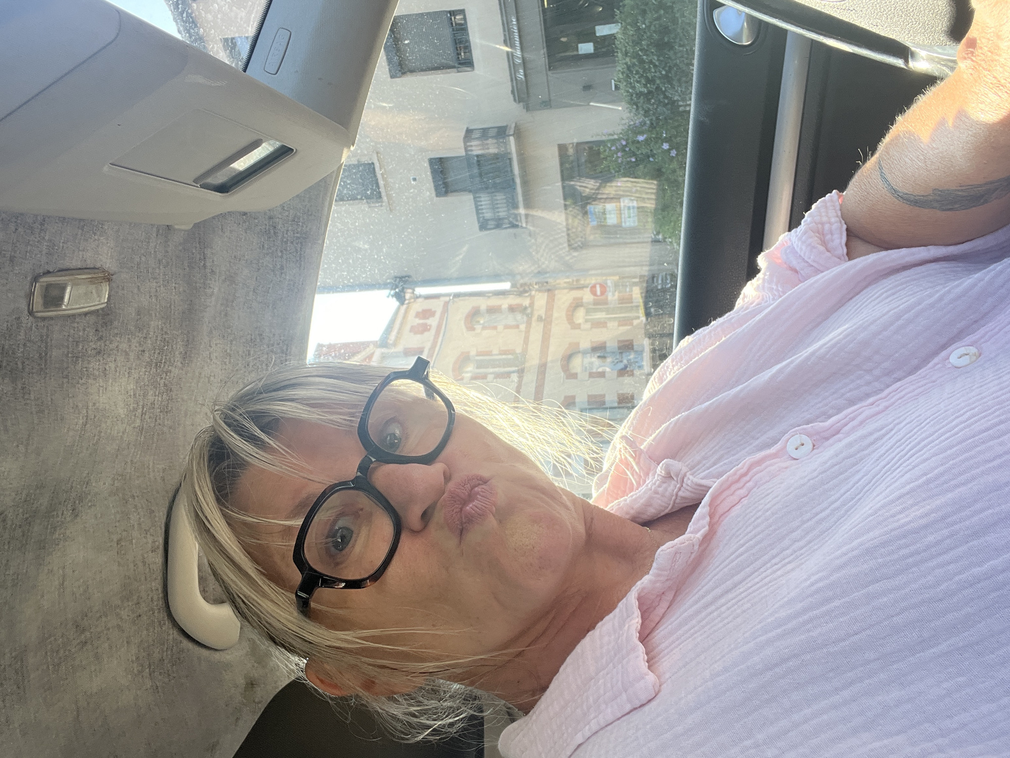 A woman is sitting in the driver's seat of a car, looking at the camera with her lips pursed. She is wearing glasses and a pink, crinkled button-down shirt. Through the car window, buildings and greenery are visible, suggesting an outdoor urban setting. The ambient lighting indicates it is daytime, possibly late afternoon due to the warm glow. The background appears to be a street scene in a town, with buildings and a hint of foliage.