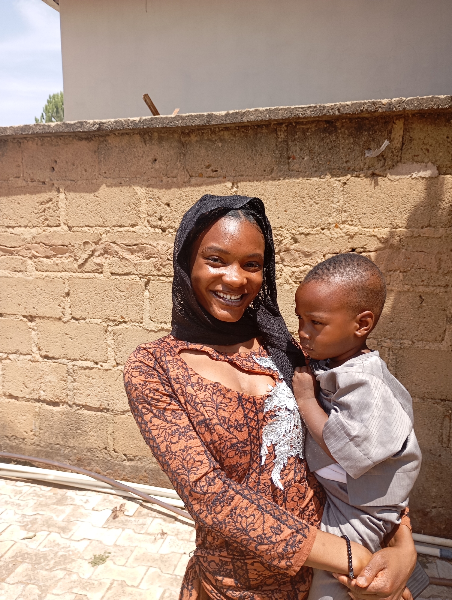 A medium shot depicts a young woman holding a small child, both standing in front of a brick wall. The woman has a broad smile, dark hair covered by a black lacy headscarf, and wears a brown patterned dress with a cutout neckline. She has a beaded bracelet on her left wrist. The child is dressed in a gray, long-sleeved outfit and appears to be looking away. The location appears to be outdoors, judging by the sunlight and brick wall background with some pipes in view. The city of Jos, Nigeria is included as the location of this photo.