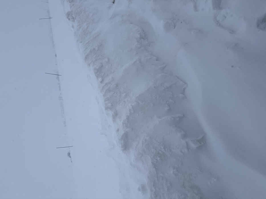 This image captures a desolate, snow-covered landscape in Kuchak, Armenia, under conditions of extremely low visibility. The foreground features a substantial snowdrift, characterized by a textured, irregular surface and sharp crests, suggesting significant wind-blown accumulation. This snowdrift extends across the lower portion and up the right side of the frame. Beyond this, the midground presents a flat expanse of undisturbed snow where two slender, dark vertical poles are visible, spaced horizontally. The background merges indistinctly with the sky, forming a uniform light grey-white, indicating heavy snowfall or dense fog. Faint, dark, linear elements are minimally discernible on the distant horizon. No individuals, animals, or vehicles are present in the scene.