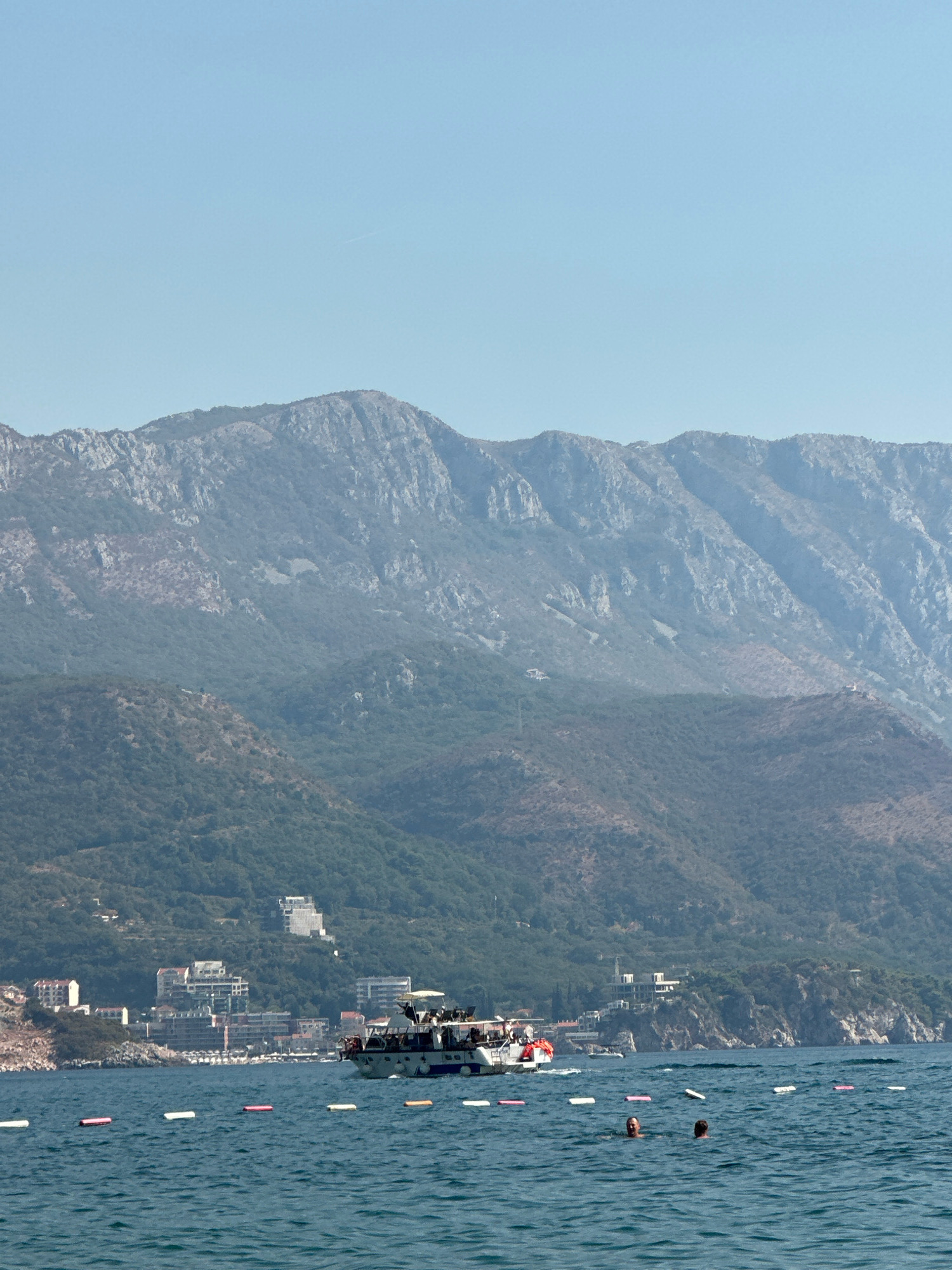 The image captures a scenic view of the Adriatic Sea with a town nestled at the foot of a mountain range. In the foreground, the blue water is dotted with buoys, and two people are visible swimming. A boat carrying passengers cruises in the middle ground, approaching the shoreline. The town of Boreti, Montenegro, is visible on the coast, with buildings clustered along the waterfront and extending up the verdant hillsides. The majestic mountain range forms a dramatic backdrop, its rugged peaks softened by a light haze under a clear, pale blue sky. The overall scene conveys a sense of tranquility and vacation, with the natural beauty of the landscape dominating the composition.