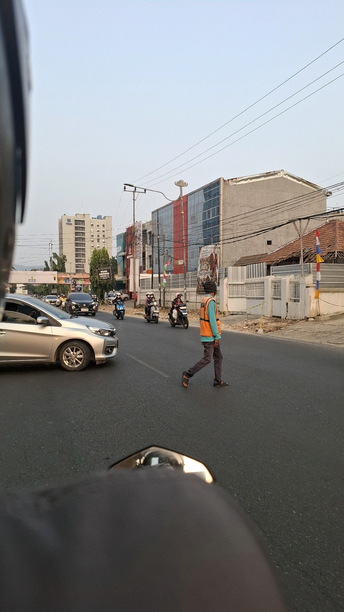 This is a photograph taken from the perspective of someone on a motorcycle, looking forward and slightly to the right. The scene depicts a street intersection in Bandar Lampung, Indonesia. On the right side of the road, a person wearing an orange safety vest and a dark hat is walking across a pedestrian crossing. They are wearing dark pants and sandals. Several motorcycles are visible on the road, with some riders and passengers. In the background, there are multi-story buildings, including a tall, light-colored building with many windows and a modern-looking building with red and blue accents. A street light is visible on the left side of the road, with power lines stretching across the frame. A silver car is also present, stopped at the intersection. The sky is clear and a light blue. The overall environment appears to be an urban street scene during daylight.