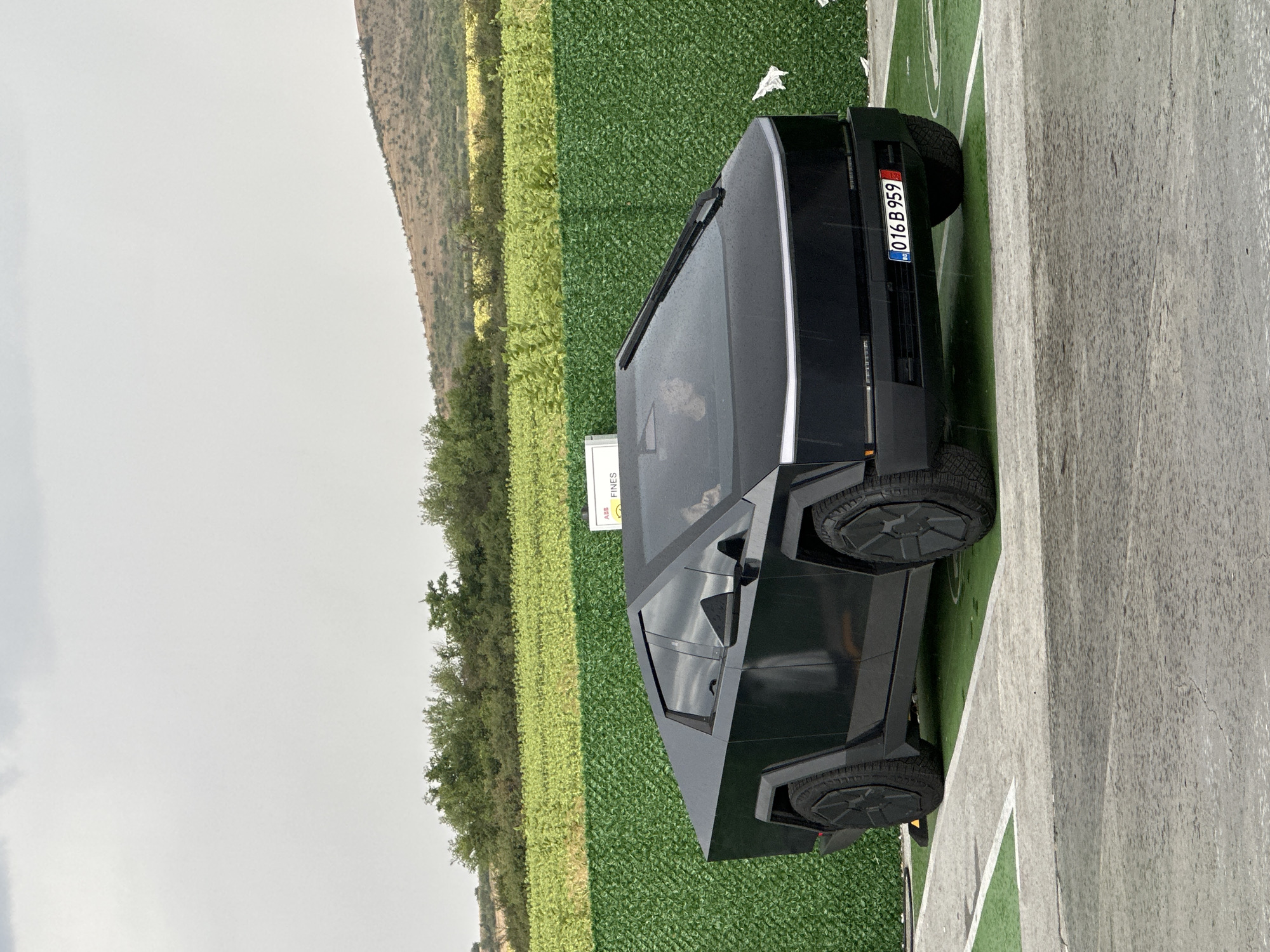 A black Tesla Cybertruck is parked in an outdoor lot, likely in the city of Polsko Padarevo, Bulgaria. The truck is angled towards the viewer's right. The sky is overcast. In the background, a green, netted fence separates the lot from a field of crops and a gentle, rolling hill. The lot itself appears to be paved with asphalt, with some sections marked with green turf and white lines, resembling a parking space. A white sign with "ASS FINES" is visible on the right side of the truck. The license plate of the truck reads "BG 016 B 959". Two people are visible inside the vehicle's cabin, though their features are indistinct. The overall atmosphere is subdued due to the cloudy weather and the stark design of the vehicle.