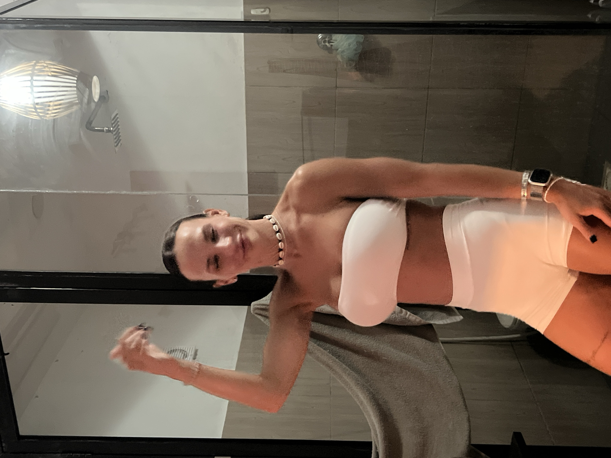 A woman in a white bandeau top and white shorts stands in a bathroom. She has dark hair pulled back and is wearing a seashell necklace. Her right arm is raised. A grey towel hangs on a rack to her left. The bathroom has tiled walls, a showerhead, and a light fixture on the ceiling. The scene is illuminated by artificial light.