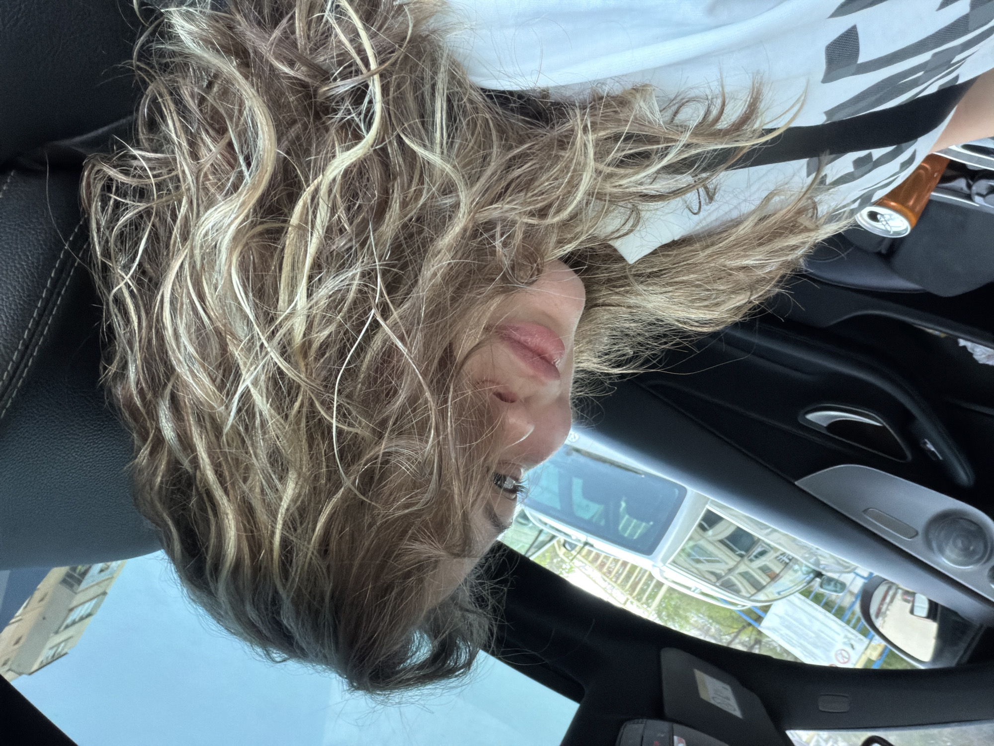 A woman with curly blonde hair is inside a car in Varna, Bulgaria. The car is parked on a city street. The view outside the car includes buildings, fencing, and signage. An open can is in the car.