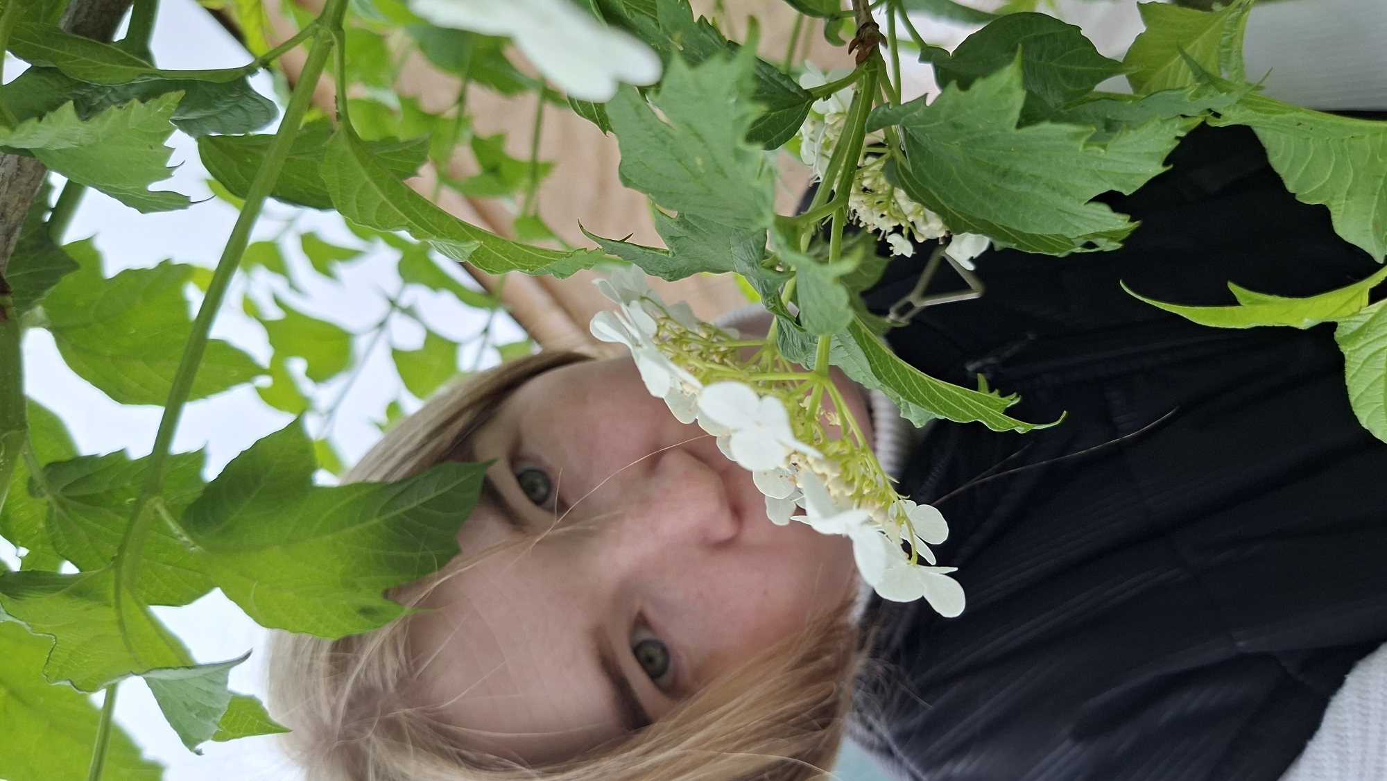 A woman with light skin, blonde hair, and green eyes is captured in a low-angle, upward-facing shot. Her face is partially obscured by green foliage and white flowers. She is wearing a dark-colored jacket with a white collar or underlayer visible. The location is speculated to be Большой Букор, Russia, but no specific location within the city can be determined based on the image. The image focuses primarily on the woman and the surrounding plants. There is no visible action or interaction occurring.
