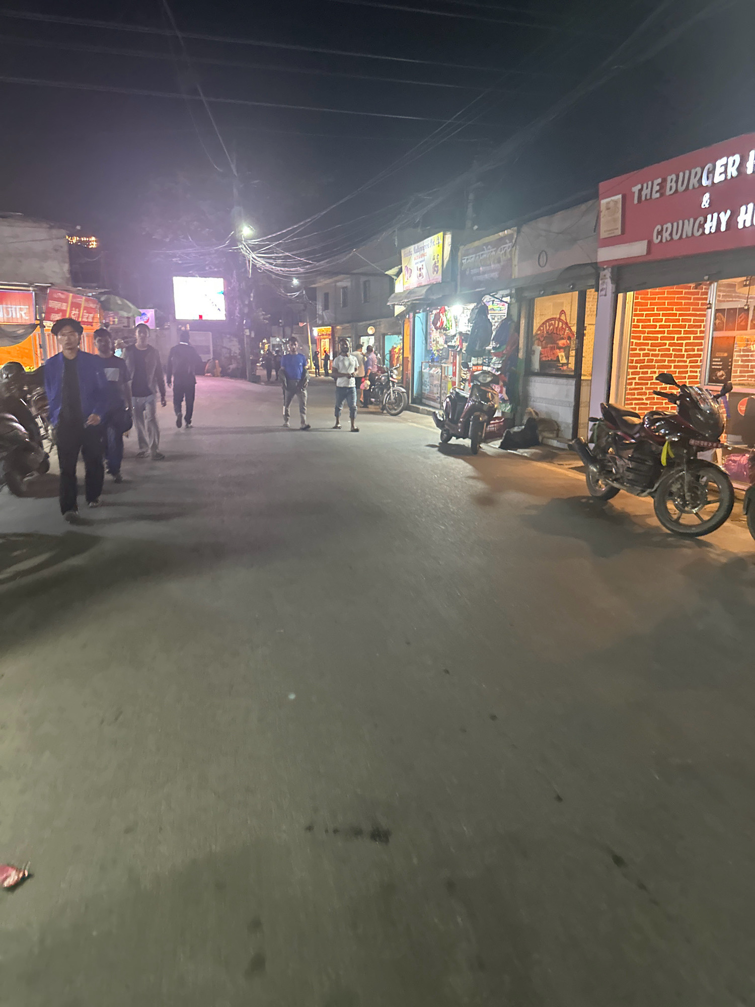 The image captures a nighttime scene on a paved street in Lalitpur, Nepal, likely a commercial area. Several individuals are walking on the street, some appearing to be locals. On the right side of the street, a restaurant called "THE BURGER H & CRUNCHY H" is illuminated, with red brick facade and glass windows displaying food items. Motorbikes are parked along the street and in front of shops. Shops with various goods are visible on both sides of the street, with some lit up. A large, bright screen is mounted on a pole in the background, displaying what appears to be an advertisement or public information. The overall atmosphere is that of a busy, illuminated street at night.