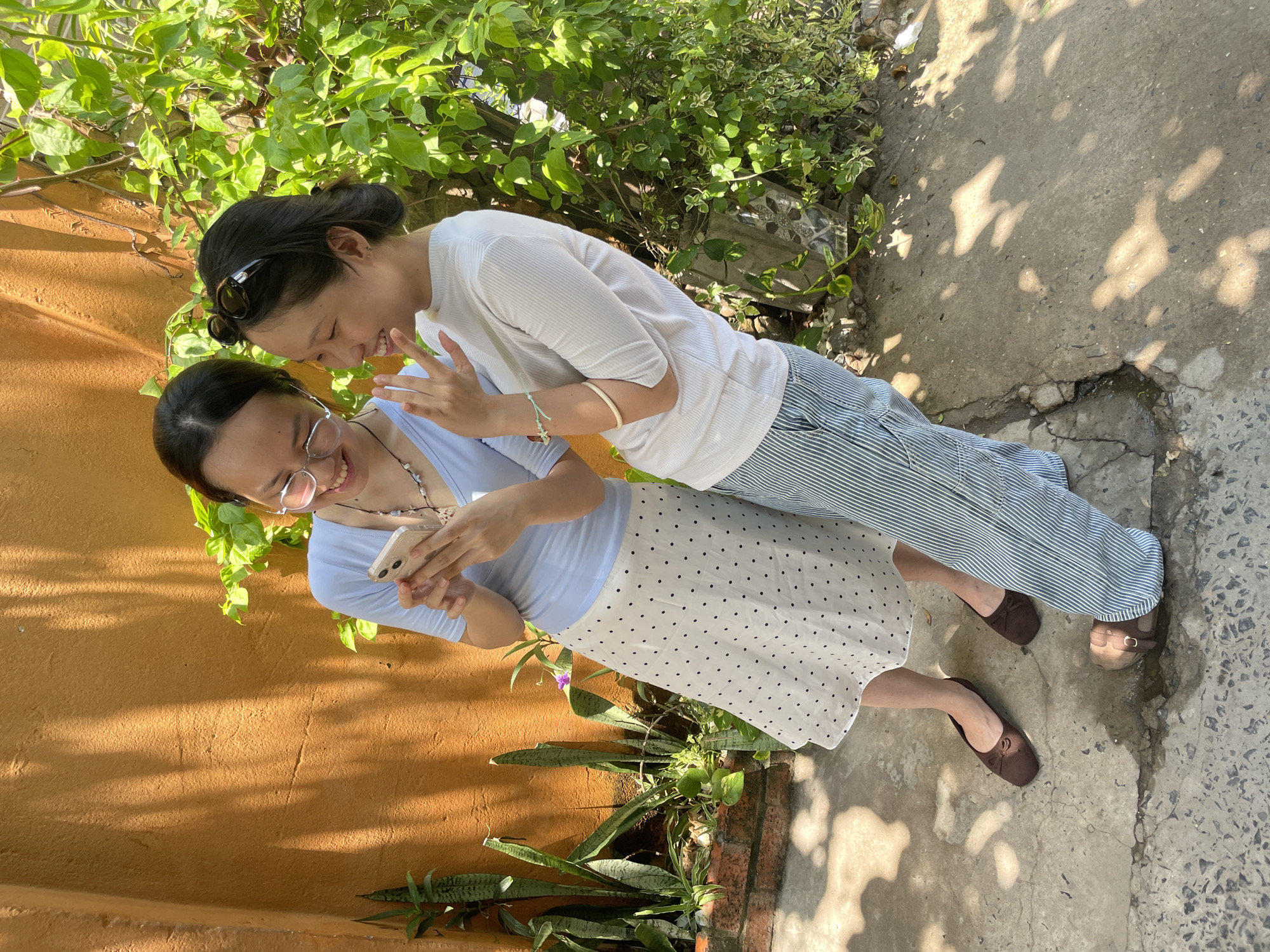 In Phan Thiết, Vietnam, two young women stand outdoors in front of an orange wall. The woman on the left wears a light blue V-neck shirt and a white skirt with black polka dots. She has glasses and is holding a smartphone, looking at it and smiling. The woman on the right wears a white ribbed t-shirt and striped green and blue pants. She has sunglasses on her head and is also smiling, looking at the phone with her hand raised. Behind them, lush green foliage grows against the orange wall, and sunlight casts dappled shadows on the concrete ground. The ground itself is cracked and shows signs of wear, with some water pooling. Both women are wearing flat shoes.