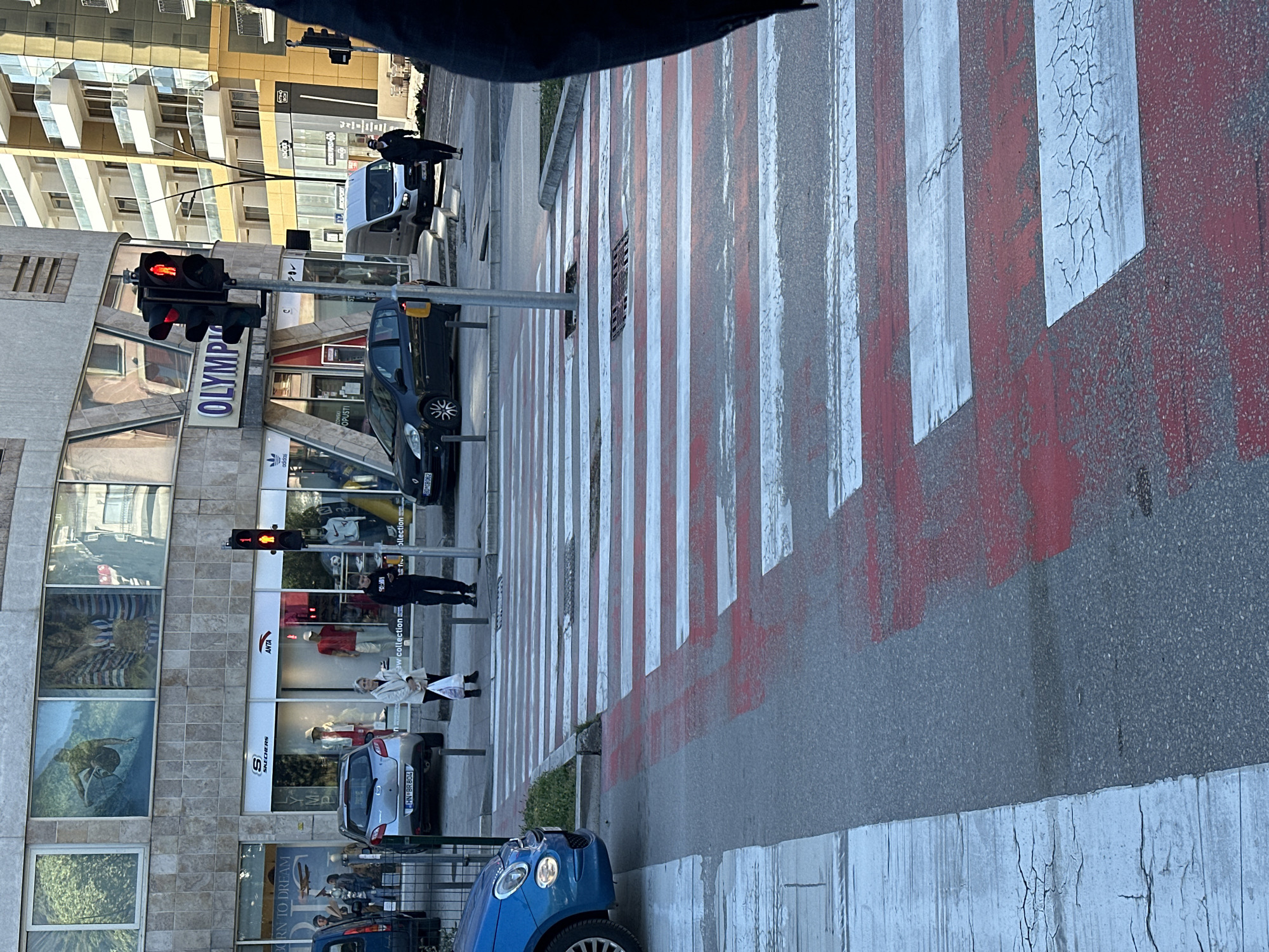The image depicts a street scene in Budva, Montenegro, featuring a pedestrian crosswalk with red and white stripes. The street is lined with buildings, including an "OLYMP" storefront and shops featuring brands like Adidas, ANTA, and Skechers. Pedestrians are visible on the sidewalk. One is near the ANTA store, another is mid-stride on the crosswalk, and a third is farther up the street. A blue vehicle and a silver vehicle are parked nearby. A black car is stopped at the crosswalk. The traffic lights in the image show a red light, indicating that pedestrians should not cross. The image is taken from a slightly elevated perspective. The bottom part of the image is darker, which could be the inside of the vehicle.