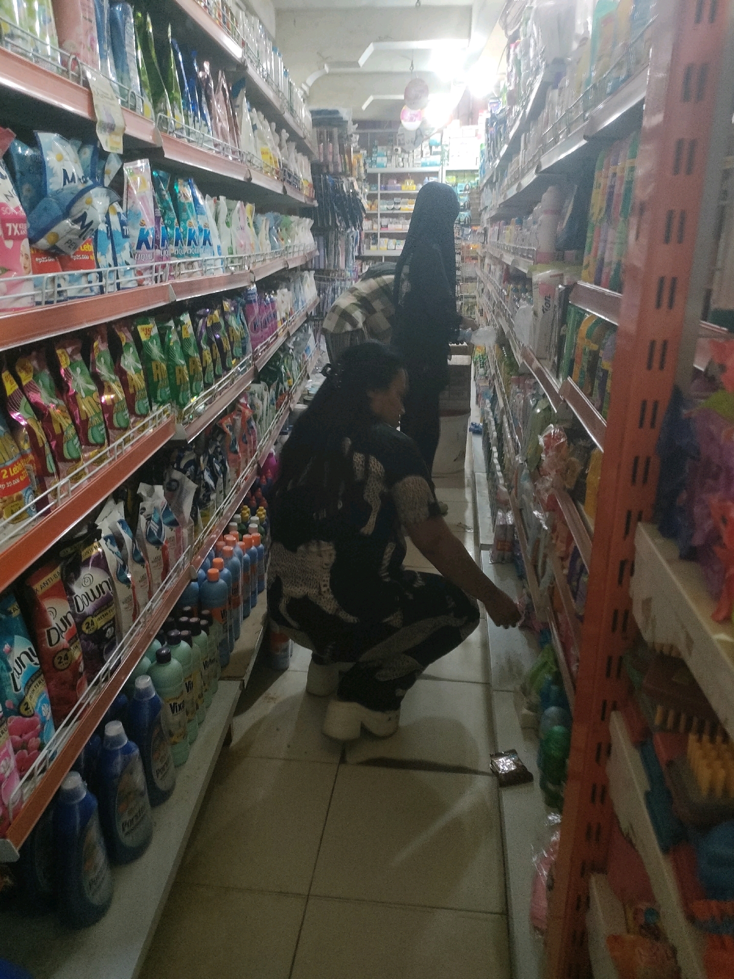 The image depicts the interior of a supermarket, specifically in the city of Salo Mallori, Indonesia. The scene is dominated by tall shelves stocked with various household products. In the foreground, two individuals are present. One, wearing a black headscarf and a long black garment, is partially visible on the left side of the image, facing away from the camera. The second individual, a woman in a black outfit with a white patterned print on the back, is crouched down on the right side, reaching for something on a lower shelf. She is wearing white shoes. The shelves are filled with a variety of items, including laundry detergents, cleaning supplies, and packaged goods. The lighting appears to be artificial, casting some shadows on the tiled floor. The overall environment suggests a typical retail setting.